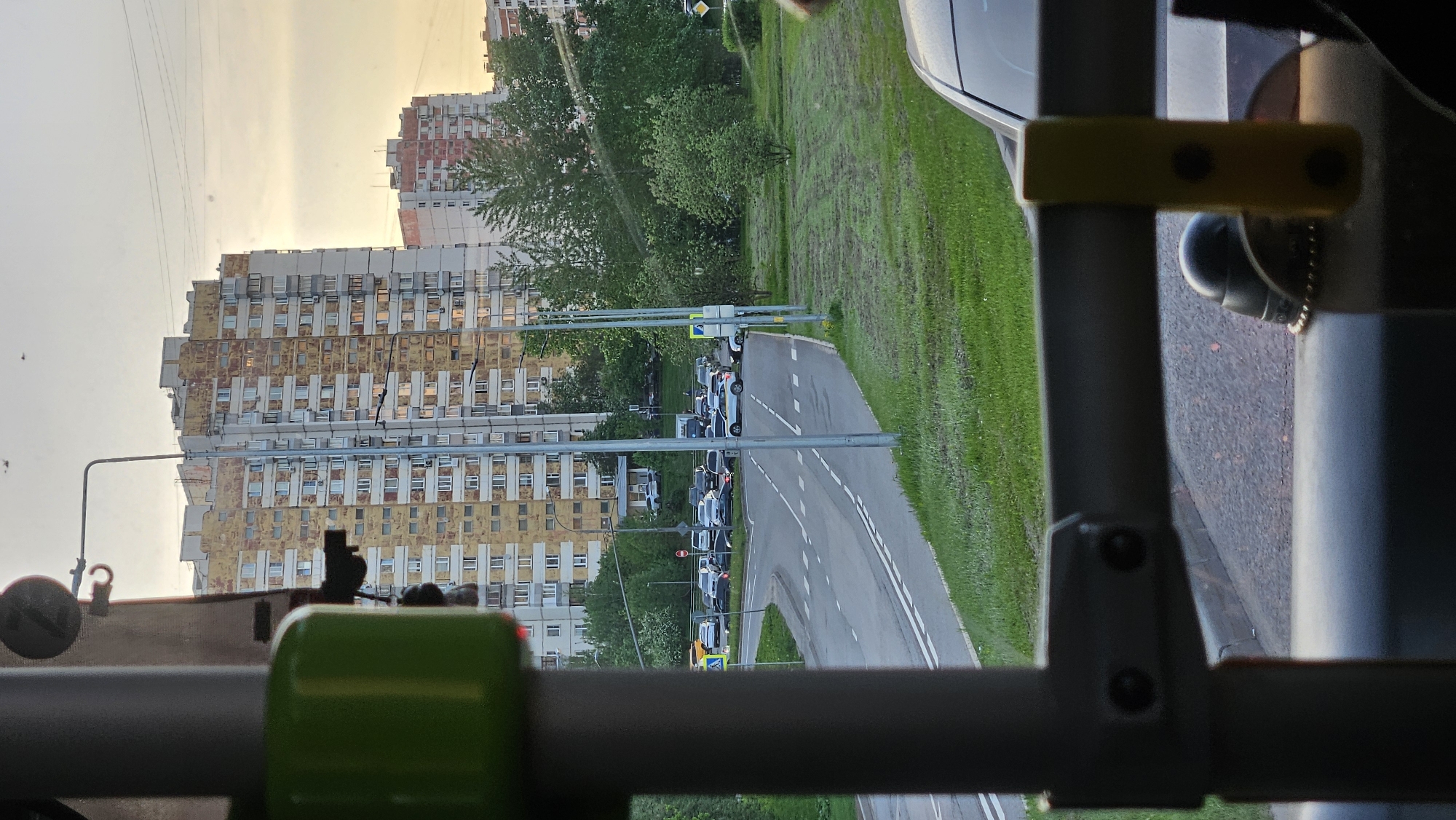 The image shows a view from inside a vehicle, likely a bus or similar transport, looking out onto a street and a block of high-rise apartment buildings. The buildings are tall, with a repeating pattern of windows, and appear to be made of prefabricated concrete panels. The buildings are showing their age with some discoloration on the exterior. The sky visible behind the buildings suggests it is either early morning or late evening due to the yellow tint.
A road runs alongside the buildings with cars parked along the roadside. The road itself has white lane markings.
The view is partially obstructed by the interior structure of the vehicle, which includes metal tubing and plastic fixtures, indicative of passenger seating or support structures. A section of the vehicle's exterior is also visible.
The scene appears to be in an urban residential area of an unknown city.