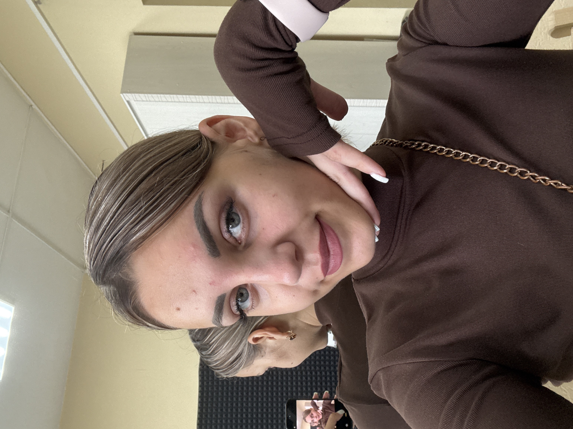 The image shows a young woman taking a selfie in a room with a light-colored wall and ceiling. She has blonde hair with highlights, blue eyes, and is wearing a brown turtleneck sweater. She is touching her chin with her right hand, which has white nail polish. A reflection of the woman and her phone is visible in a mirror behind her. The room appears to be an indoor space, possibly a studio or a dressing room, with some soundproofing panels on the wall. There is also a piece of furniture that looks like a wardrobe or cabinet in the background. There is no indication of the specific location being Yuzhno-Sakhalinsk, Russia.