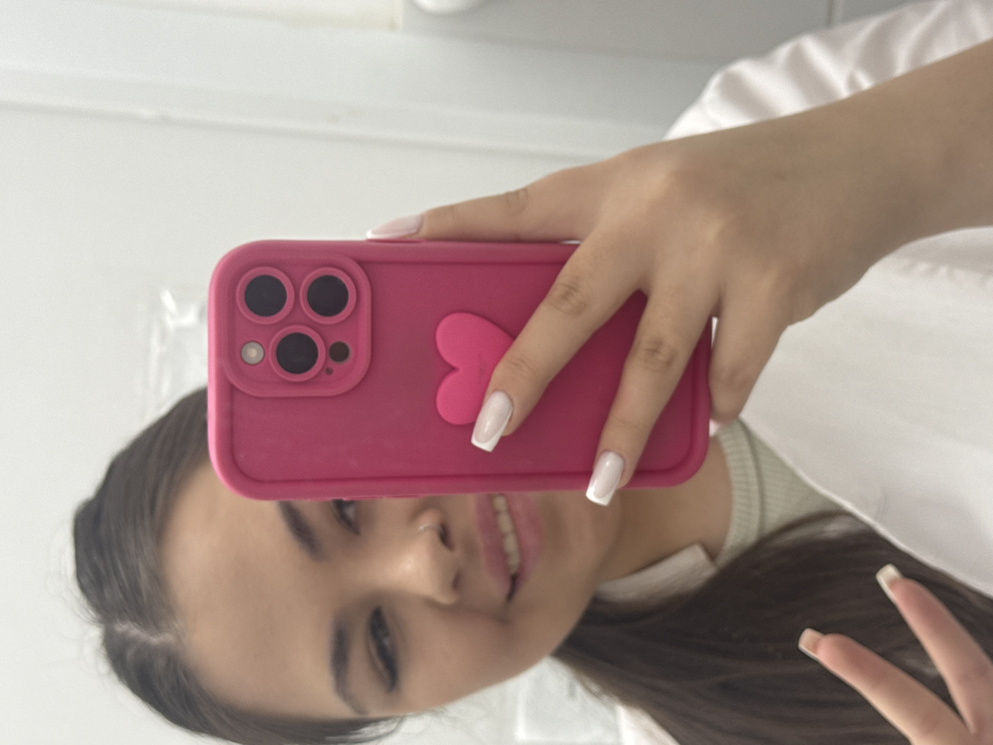 A woman is taking a selfie in what appears to be an indoor setting. She is holding a pink smartphone with a heart-shaped decoration on the back. Her hair is dark, and her nails are manicured with white tips. She seems to be smiling slightly. The background is blurry, but it suggests a clean, possibly clinical, environment. The location is unknown, and no specific elements indicate the city of Pargolovo, Russia.