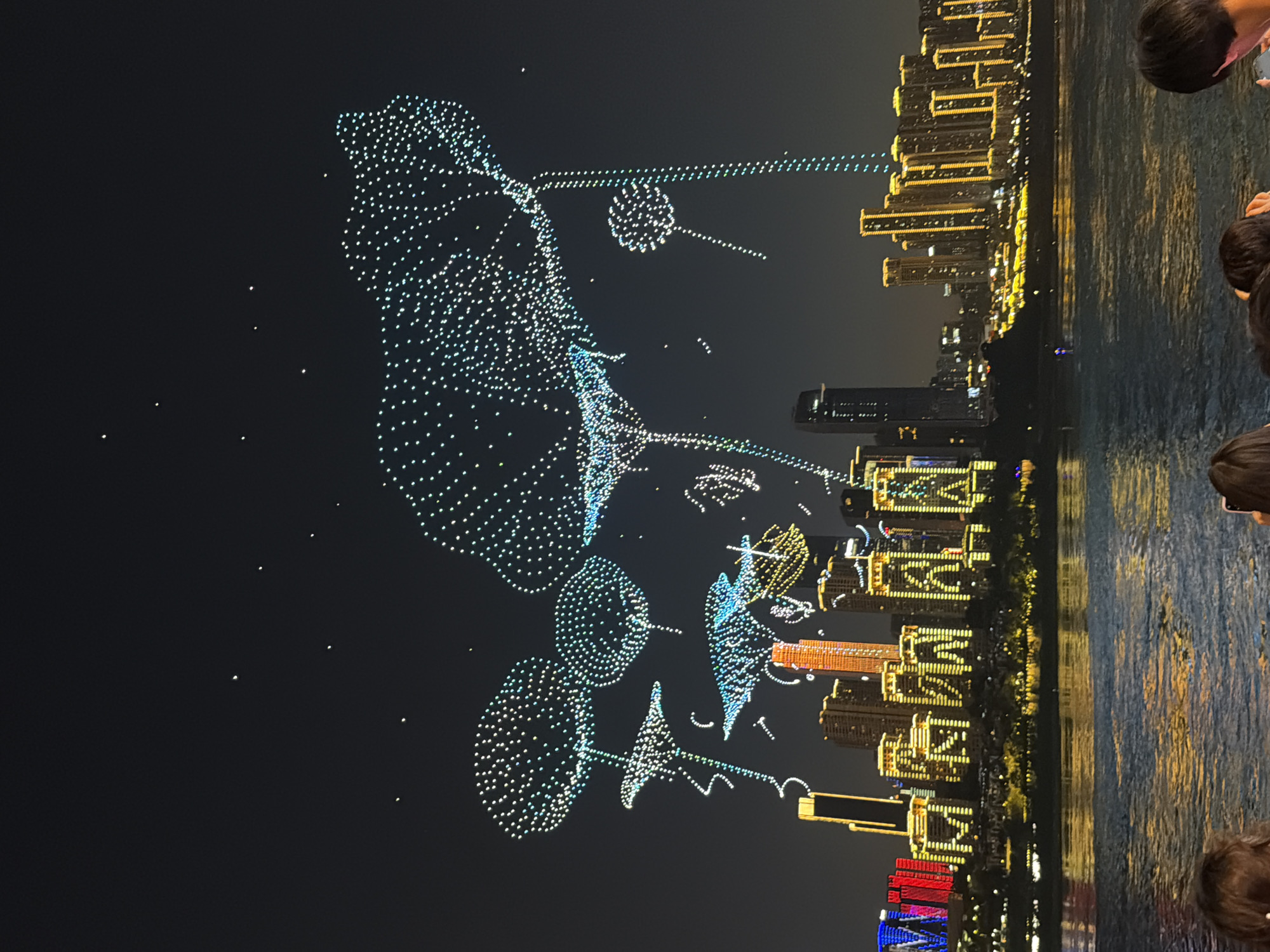 This image captures a nighttime drone light show over the Nan'an District in China. The sky is filled with a complex formation of numerous small lights, forming abstract shapes and figures that appear to be in motion. These illuminated structures are suspended high above a cityscape characterized by numerous tall buildings. The buildings themselves are illuminated with yellow and white lights, outlining their architectural features and creating a vibrant urban backdrop. In the foreground, a body of water reflects the lights from the city and the drone formations, creating a shimmering effect on the surface. The bottom edge of the frame shows the heads and shoulders of several people, suggesting they are spectators observing the event from a vantage point near the water. The overall scene is one of a modern urban environment illuminated by a dazzling display of technology and light.