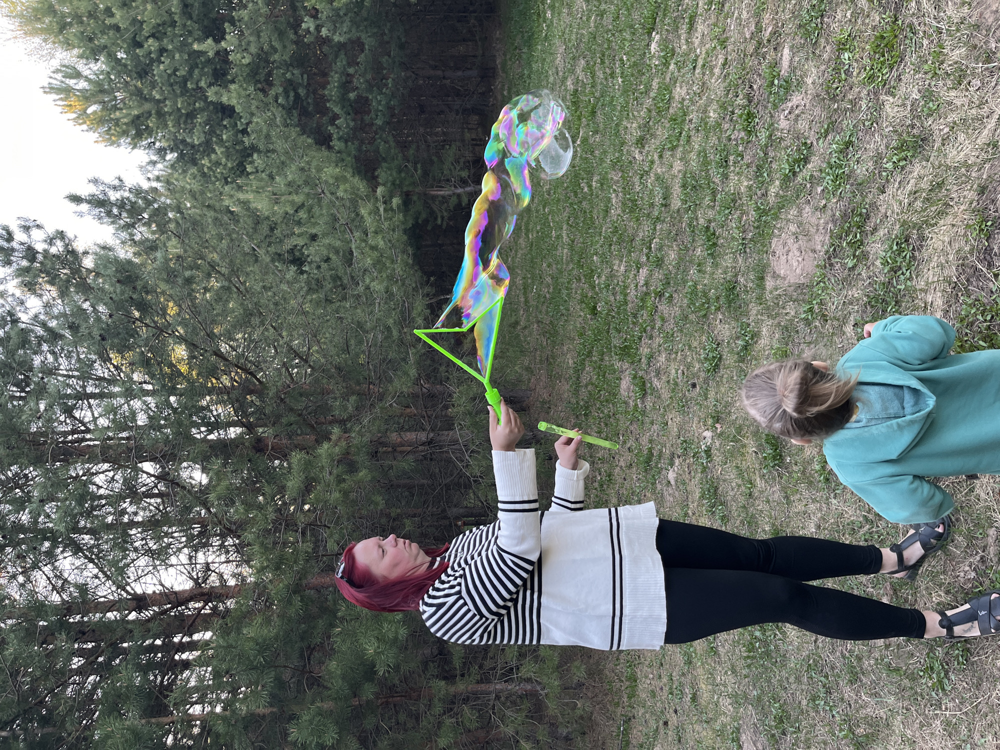 The image depicts an outdoor scene, likely in a park or green space, within the city of Chaikovsky, Russia. A woman with reddish-purple hair is seen holding a bubble wand, from which large, iridescent bubbles are being created. She is wearing a black and white striped sweater, black leggings, and sandals. Beside her, a child with blonde hair, wearing a green hooded sweatshirt, appears to be watching the bubbles. The background consists of trees and grassy terrain.