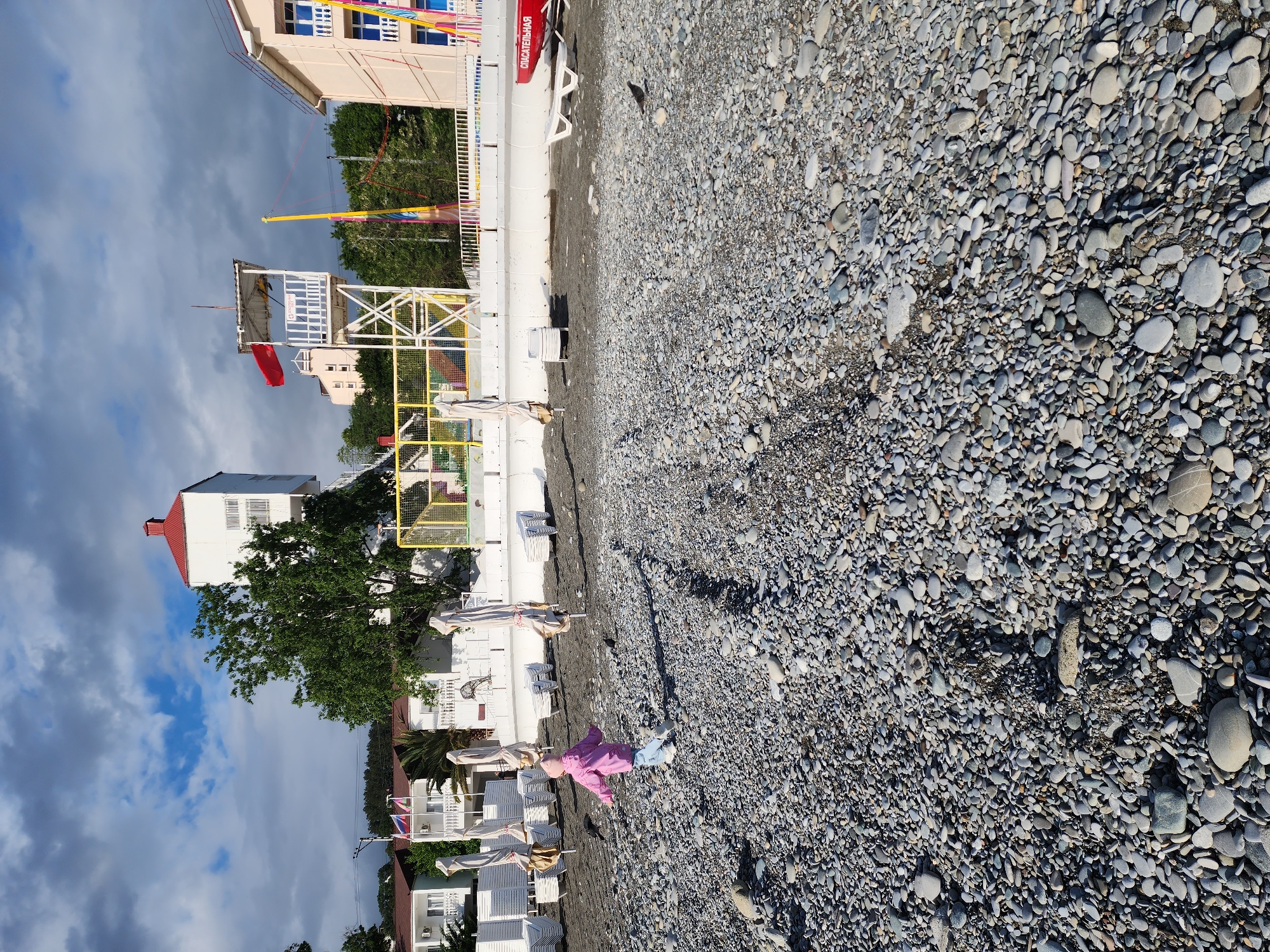 A beach scene at Sochi, Russia. The ground is covered in dark grey stones. A girl in a pink jacket and blue pants lies on the stones. Beach infrastructure lines the back of the beach, including white sun umbrellas, white buildings, a lifeguard tower, and a trampoline attraction. Overcast sky.