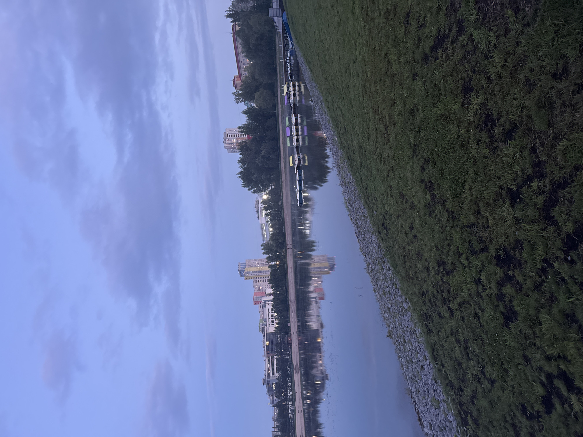 This is a landscape photograph taken at dusk on a lake, likely in the city of Ufa, Russia. In the background, a skyline of multi-story buildings and trees is visible, with reflections on the calm water surface. Along the edge of the water, a row of colorful paddle boats are docked, some yellow, purple, and green. The foreground consists of a grassy slope leading down to a rocky embankment along the lake's edge. The sky is cloudy and a soft blue, indicating the end of daylight. There are no people clearly visible in the image. The scene depicts a peaceful recreational area by the water.