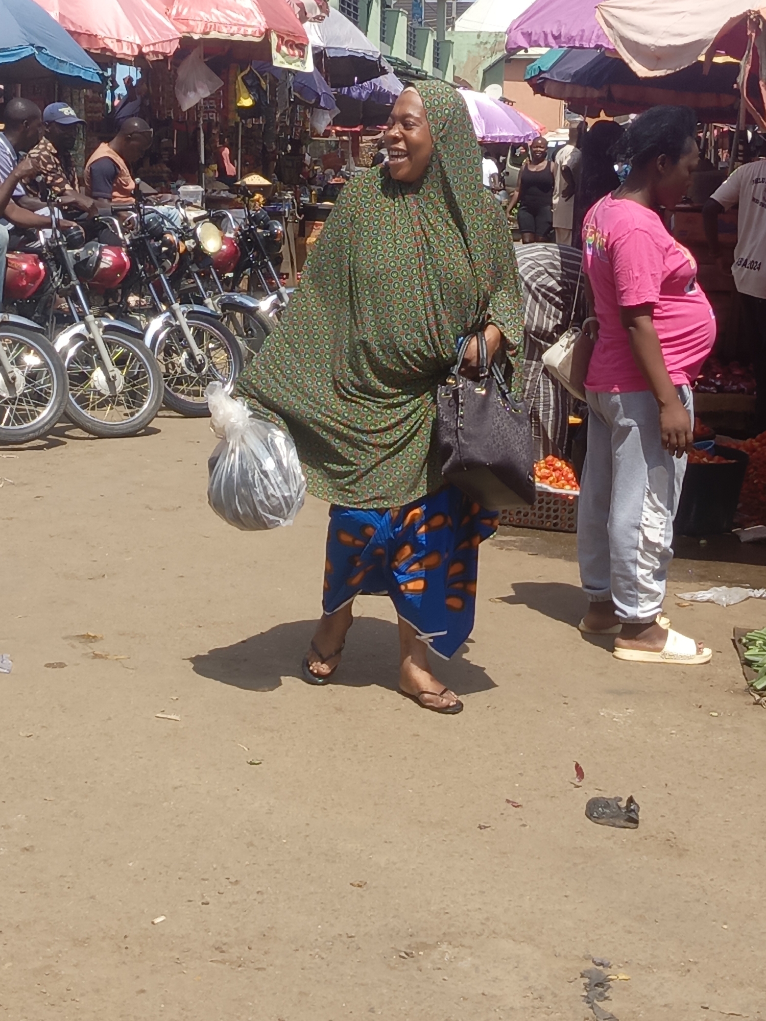 An outdoor market scene in Akwanga, Nigeria during daylight. The ground is a sandy brown surface. A woman with a green patterned head covering and tunic over a blue patterned skirt walks, smiling, while carrying a black purse on her shoulder and a plastic bag filled with dark items in her right hand. Next to her stands a pregnant woman in a pink shirt and gray pants. Several motorcycles are parked nearby. Vendors and shoppers are present, obscured by market stalls and umbrellas in the background.