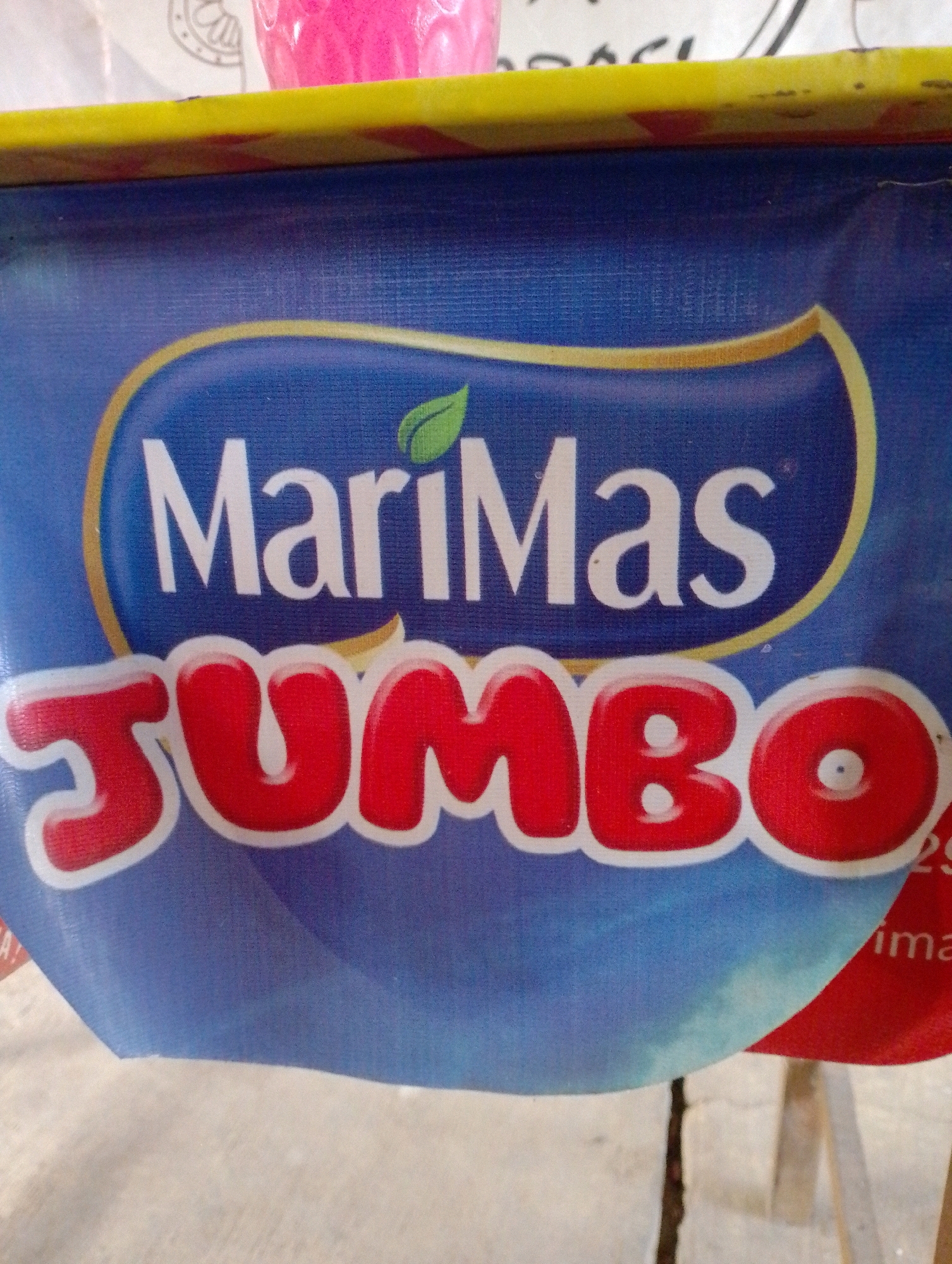 The image shows a close-up of a blue banner with a yellow border at the top. The banner features the text "MariMas Jumbo" in white and red, with a green leaf graphic. The banner appears to be part of a display or advertisement. In the background, a blurred pink object is visible, possibly a cup or container. The lower part of the image shows a textured surface that could be a table or counter. The context of this image is from a street food stall in Ngeling, Indonesia, indicated by the banner's branding and the typical setting of such stalls.