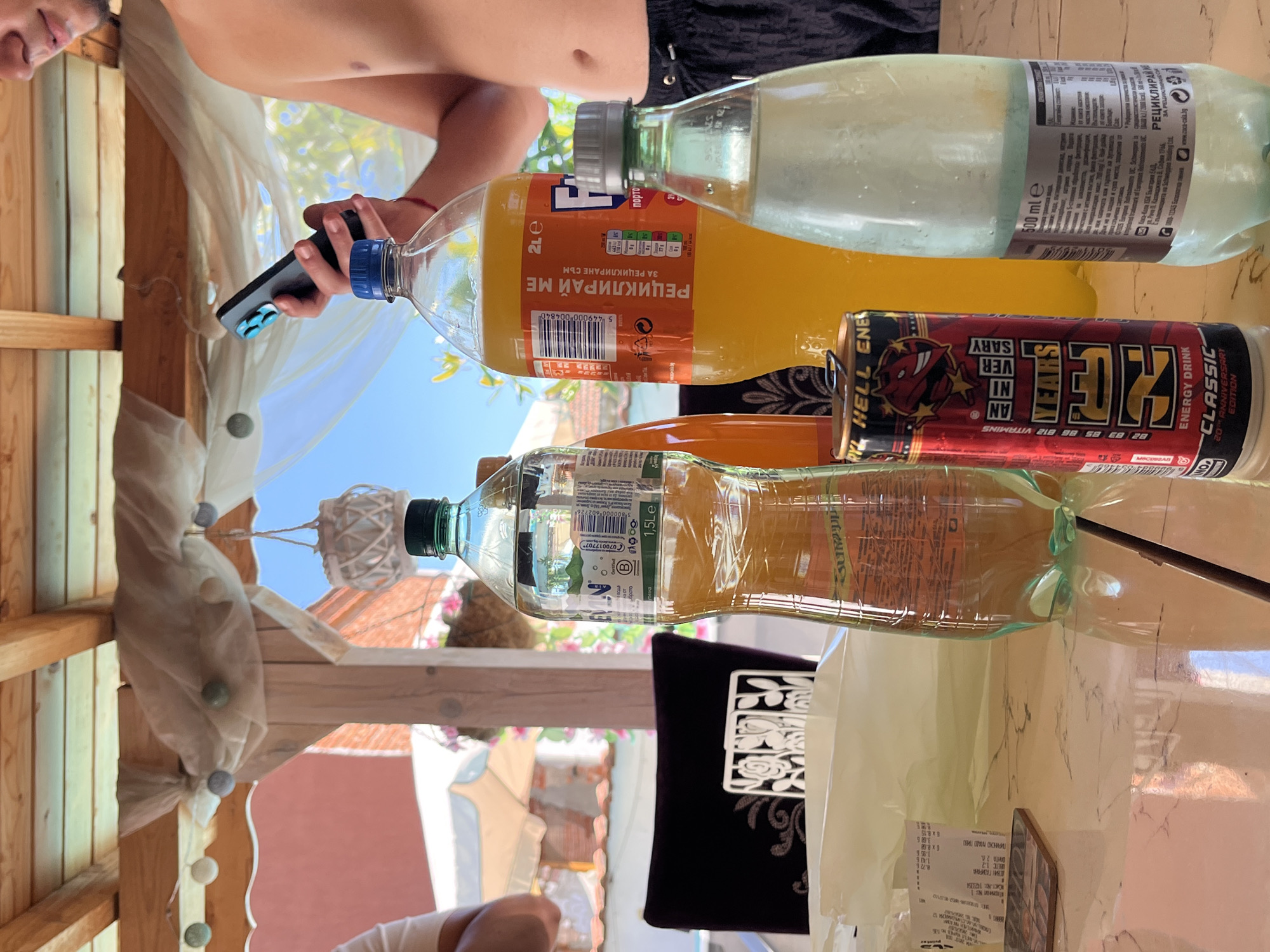 Here is a forensic description of the image:
The image shows an outdoor table scene with several beverage containers. Present are two plastic bottles of clear soda, one labeled "1.5Le" and the other "500 ml e". There is also a 2-liter bottle of Fanta orange soda, and a can of Hell Energy Drink Classic 20th Anniversary Edition. A receipt is also visible on the table. A person is holding a mobile phone in their hand, partially visible at the top-left corner of the image. Another person is partially visible in the lower portion of the image. The table is under an awning and the backdrop reveals a partially visible cityscape. It is highly probable that it is in Elhovo, Bulgaria based on the language on the beverage containers.