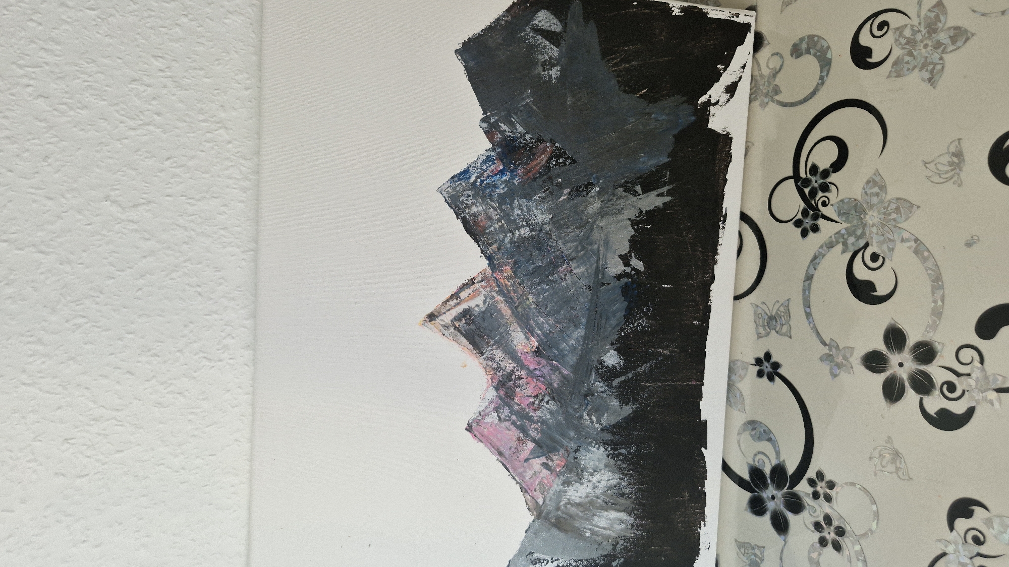 The image shows a painting of mountains on a white canvas. The mountains are rendered in shades of gray, pink, and blue, with dark black at the base suggesting trees or a forest. The painting is placed on a surface with a light-colored floral pattern. The background of the image is a textured white surface, likely a wall. There is no indication of the city of Baden-Baden, Germany.