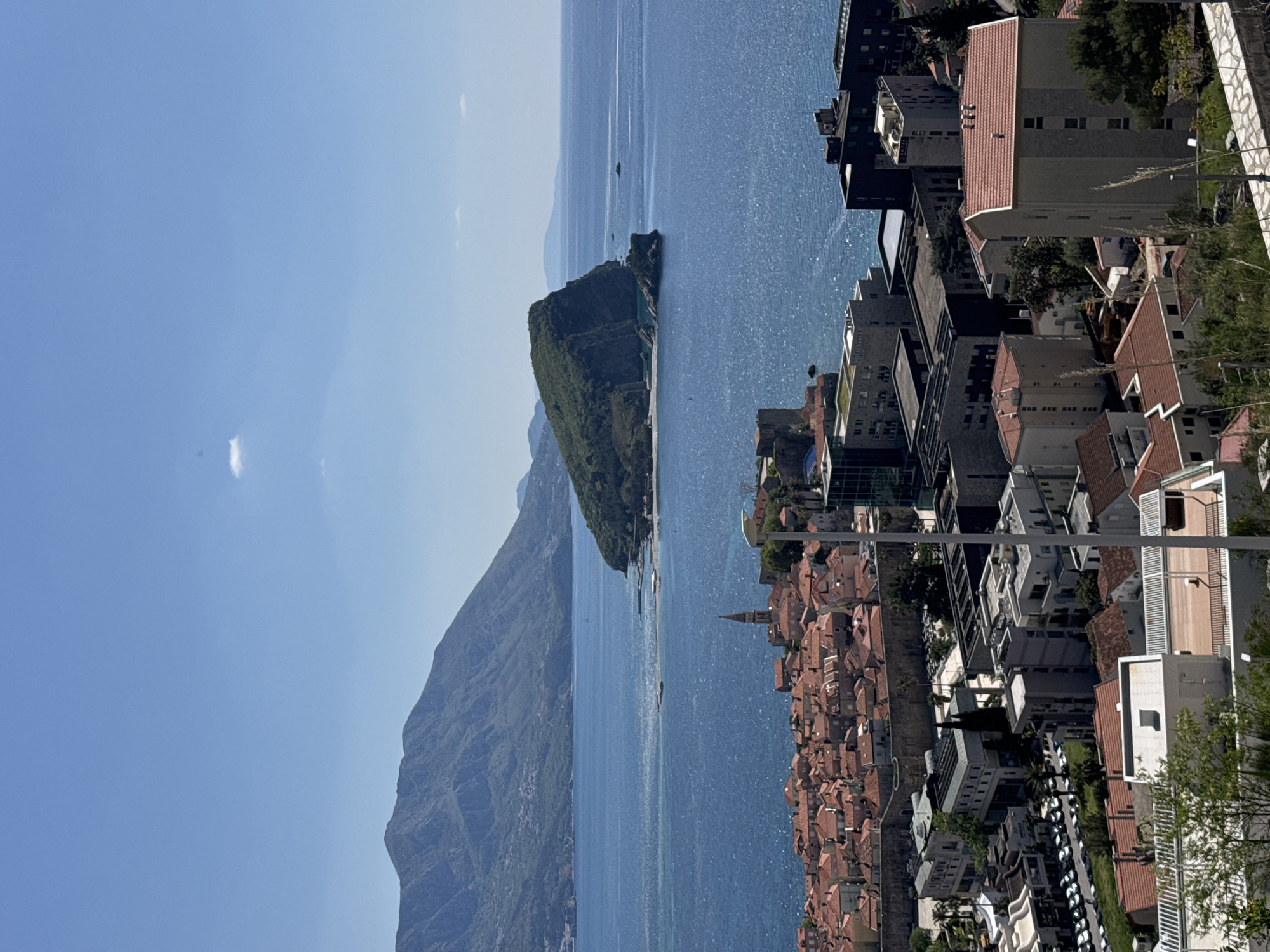 The image displays a panoramic coastal landscape from an elevated viewpoint, looking out over a wide expanse of sparkling blue sea. The sky above is a clear, bright azure, with only a few faint, wispy clouds visible.
In the midground, the sun-drenched sea shimmers intensely, reflecting light off its surface. To the left, a range of green, rugged mountains stretches along the horizon, receding into a light haze. A prominent, verdant, rocky island or peninsula extends into the sea from the left side of the frame.
On the right, a densely built-up city or town occupies the coastline and climbs up the adjacent hillside. The architecture is a mix of older buildings with distinctive red-tiled roofs, characteristic of Mediterranean or Adriatic regions, and some more modern, taller structures. A slender church bell tower or spire is visible among the red rooftops. Green trees and shrubs are scattered throughout the urban area and on the surrounding terrain.
The scene is illuminated by bright daylight, suggesting mid-morning to early afternoon, with strong sunlight creating clear visibility and reflections. No specific activities or people are discernible. Partial text, "HOTEL VAL...", can be observed on one of the buildings in the cityscape.