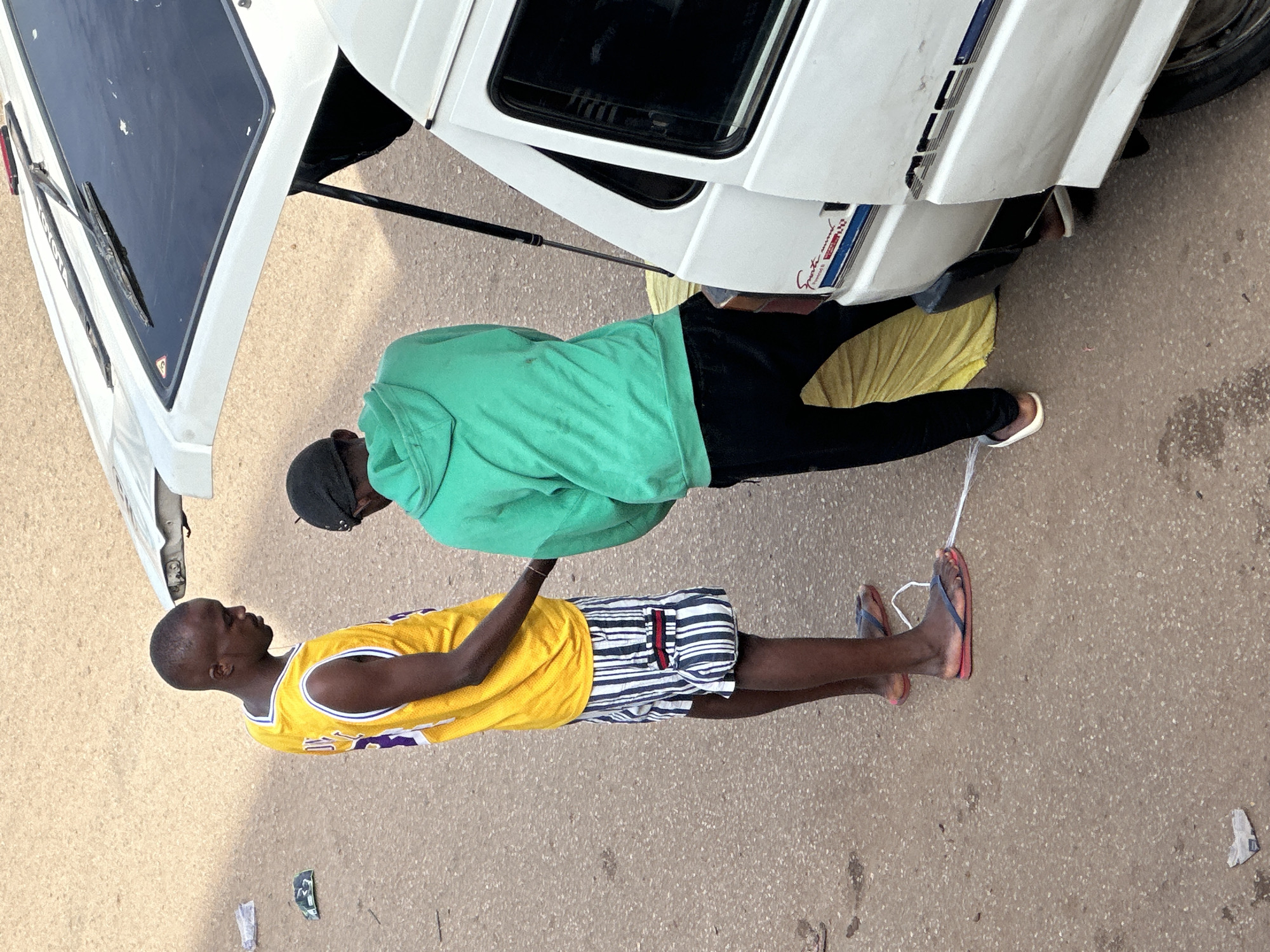 Two young men are standing next to a white van with its rear hatch open. The man on the left is wearing a yellow basketball jersey with purple trim and white and blue striped shorts. He has short hair and is looking down at the other man. The man on the right is wearing a green hooded sweatshirt and black pants. He appears to be interacting with something inside the open van. The ground is asphalt and there is some litter visible. The scene appears to be in an urban or semi-urban environment in Bukuru, Nigeria.