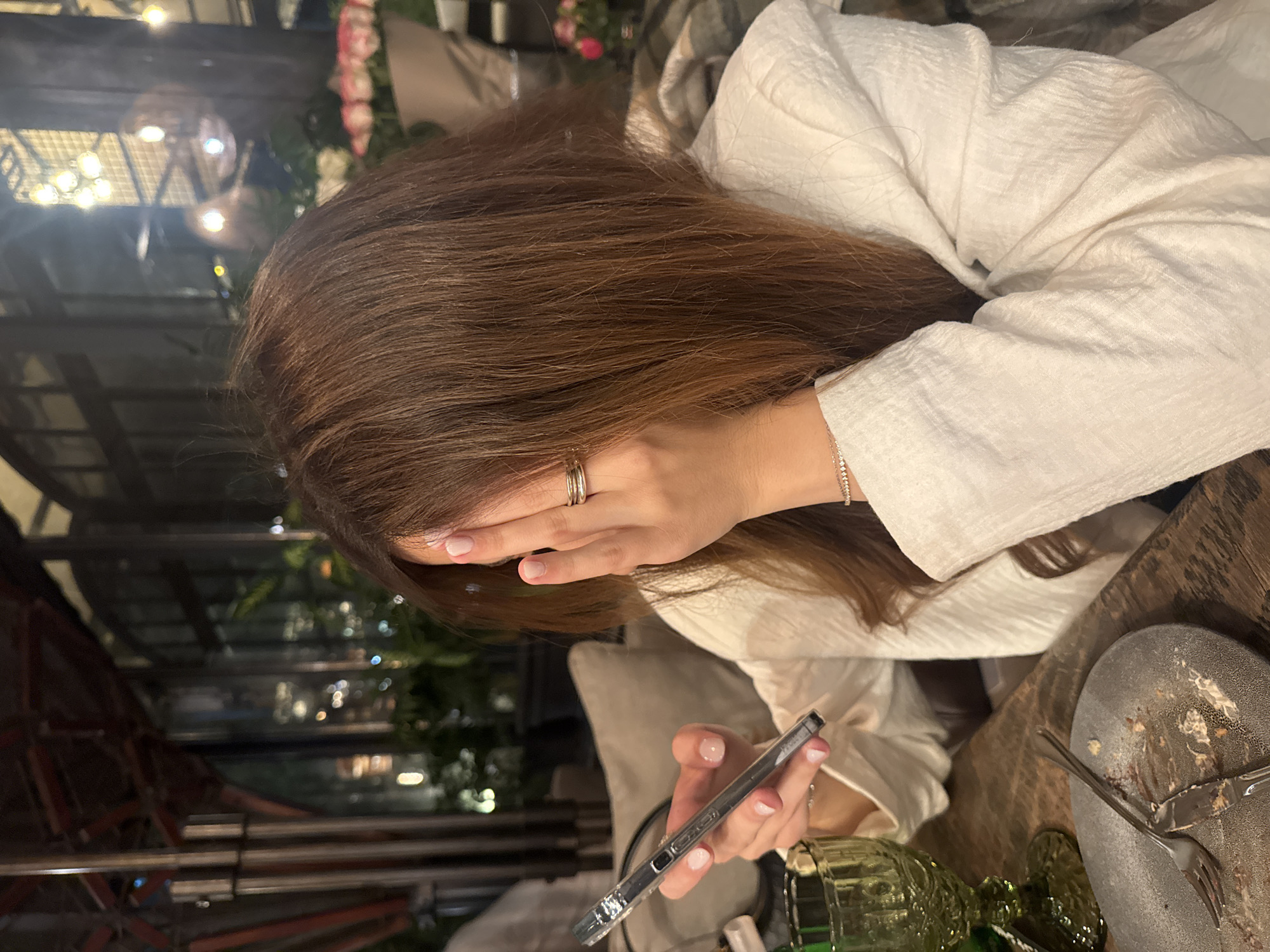 A person is seated at a table in Yerevan, Armenia, with their head partially obscured by their hair and hand. The individual is holding a smartphone in their left hand. The table setting includes a dirty plate with used cutlery, a glass, and possibly another object partly visible. The background shows a dimly lit area with blurred city lights and vegetation.