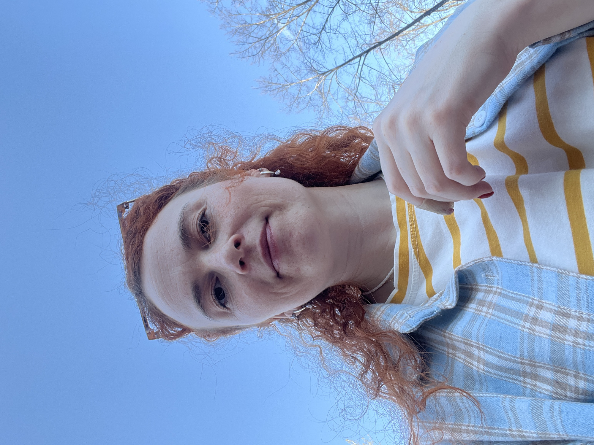 A low-angle selfie shows a woman with reddish, curly hair and fair skin. She has visible freckles on her face. She is wearing glasses perched on her hair. The woman is wearing a yellow and white striped top under a blue and white plaid shirt. Her hand is partially visible on the right side of the frame. The background is a clear blue sky with the bare branches of trees visible above her. There is no information in the image to suggest this was taken in Chaikovsky, Russia.