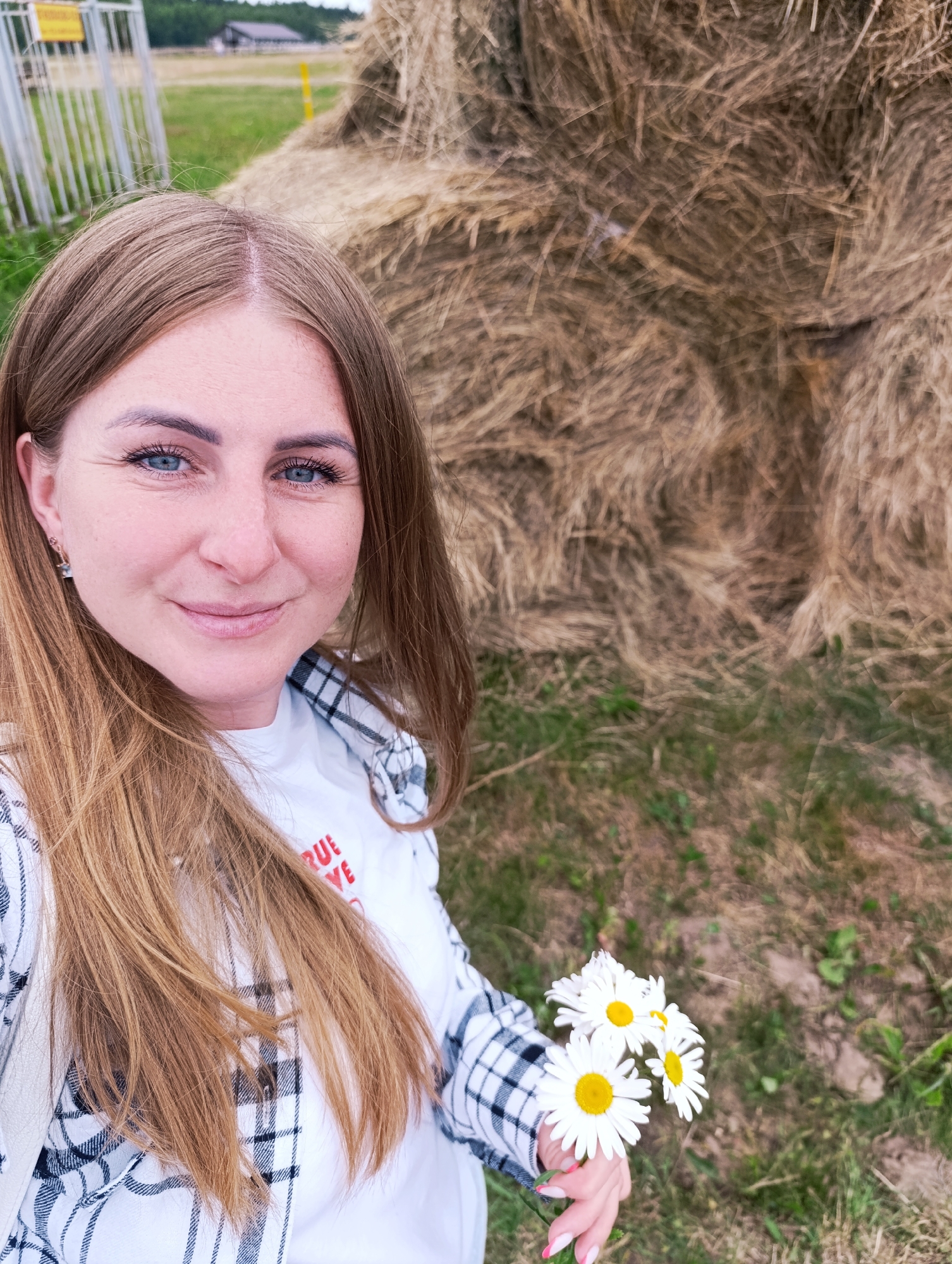 A woman with long, light brown hair and blue eyes smiles into the camera, holding a small bouquet of daisies. She is wearing a white t-shirt with red text that reads "TRUE LOVE" and a black and white checkered shirt over it. Her left hand, with bright pink painted fingernails, is visible holding the flowers. She has a small earring in her left ear.
The background consists of a large haystack, and grassy ground with some dry patches. In the far left of the image, a metal fence and a yellow sign are partially visible, suggesting an outdoor rural or agricultural setting. The lighting suggests it is daytime. The overall mood is cheerful and relaxed. The scene is set in Ivanovskoe, Russia.
