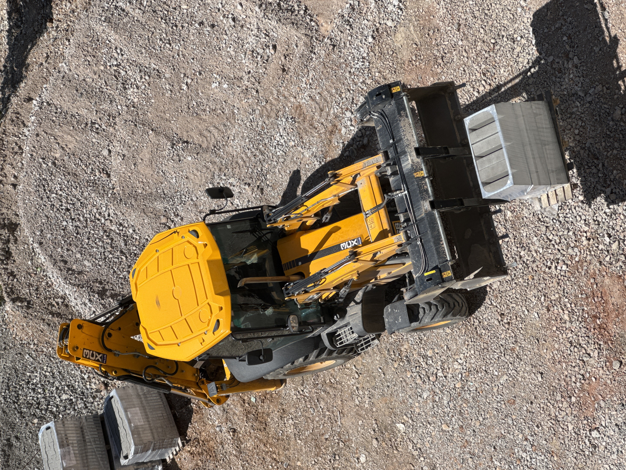 An aerial, slightly oblique view captures a yellow and black backhoe loader (digger) stationary on a gravel and dirt-covered construction site in Sofia, Bulgaria. The large machinery, with its front loader bucket and rear excavator arm, occupies the central and left portions of the frame.
The ground is a mixture of light grey and brown gravel and fine dirt, typical of an undeveloped or active construction area. Strong, sharp shadows cast by the vehicle and other objects indicate bright, sunny weather, likely during the morning or late afternoon.
On the right side of the image, next to the backhoe loader's front bucket, is a wooden pallet stacked with numerous light grey, rectangular building blocks or pavers, neatly wrapped in clear plastic. A similar stack of wrapped blocks is partially visible in the bottom-left corner of the image, near the vehicle's rear.
No people are visible in the scene. The backhoe loader appears to be currently inactive, possibly between tasks of moving materials or preparing the ground. Visible text on the yellow side panel of the vehicle clearly displays the brand name "MUX" in black lettering. Other small warning symbols are present on various black components of the machinery. The image depicts a moment of pause at a construction or development site.