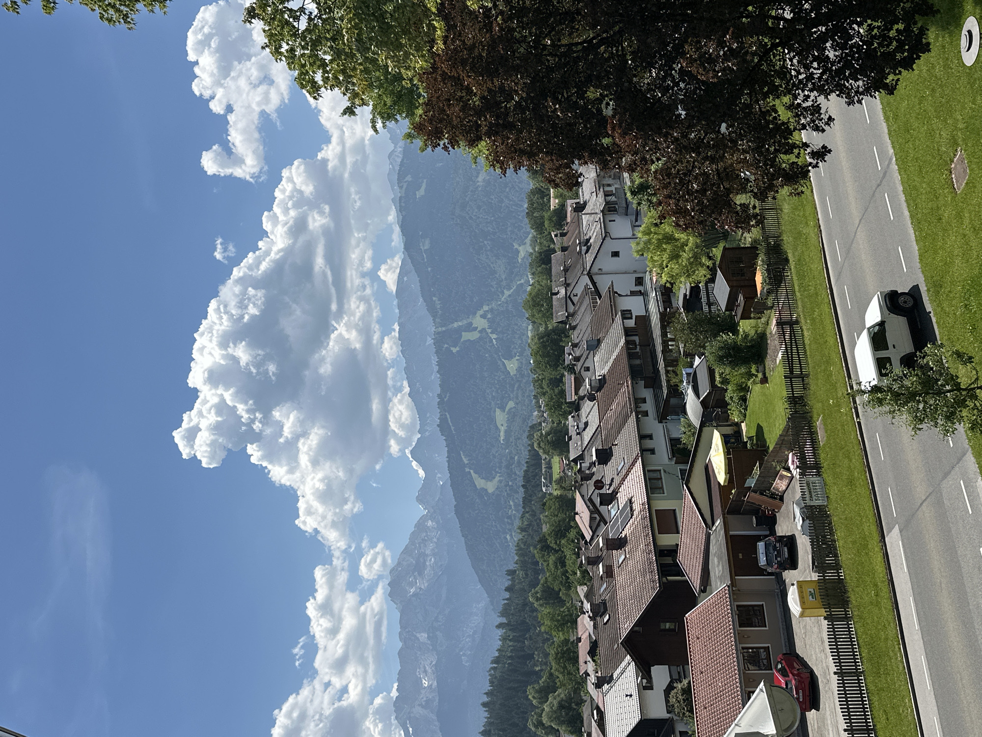 This is a high-angle, eye-level outdoor photograph taken on a clear day in Garmisch-Partenkirchen, Germany. The image captures a panoramic view of a residential area nestled against a backdrop of majestic mountains and a vibrant blue sky filled with large, fluffy white clouds.
In the foreground, a paved road runs horizontally across the frame. A white SUV is driving on the right side of the road. To the left of the road is a well-maintained green lawn. Beyond the lawn, a wooden fence separates the properties from the street.
The mid-ground of the image showcases a collection of red-tiled roof houses with white or light-colored walls, typical of European architecture. Some houses have solar panels on their roofs. A red car and a dark-colored SUV are parked in a driveway next to a small yellow portable toilet. A yellow umbrella is set up in one of the yards. Several garden sheds and fences are visible.
The background is dominated by lush green hills and dense forests. Further in the distance, a majestic mountain range with snow-capped peaks rises against the sky. Ski slopes are visible on some of the mountain faces, suggesting the area is a popular destination for winter sports. The overall atmosphere is serene and picturesque, highlighting the natural beauty of the Bavarian Alps.