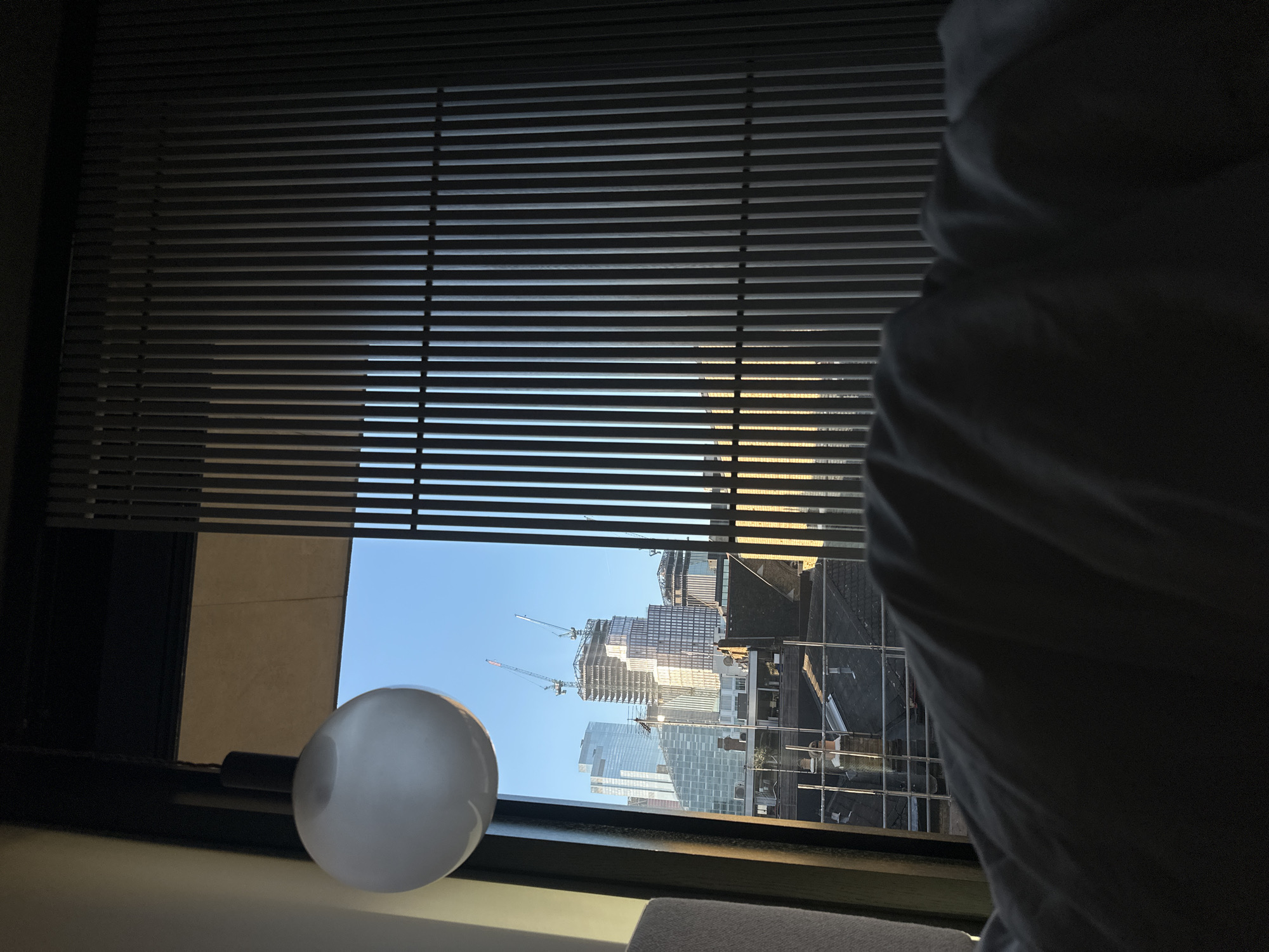 The image presents an interior vantage point looking out a window onto a cityscape in the City of London, United Kingdom, under a clear blue sky during daylight. In the upper-left, a white spherical pendant light hangs from the ceiling. The bottom of the frame is dominated by a dark, rumpled fabric, likely bedding, suggesting a bedroom setting. To the right, a series of vertical, narrow slats form a screen or blind, partially obscuring the view.
Through the window, a complex urban environment is visible. Prominent in the midground are several modern skyscrapers. On the left, a finished glass-clad building with a distinctive stepped, diagonally segmented facade is identifiable as 100 Liverpool Street. Adjacent to it, a tall building is under significant construction, featuring a visible skeletal frame and two active tower cranes at its summit, identifiable as 8 Bishopsgate. Below these towering structures, in the immediate foreground, are numerous traditional low-rise buildings characterized by dark pitched roofs, multiple chimneys, and visible scaffolding. Direct sunlight illuminates elements of the cityscape and casts distinct shadows, particularly on the slatted screen.