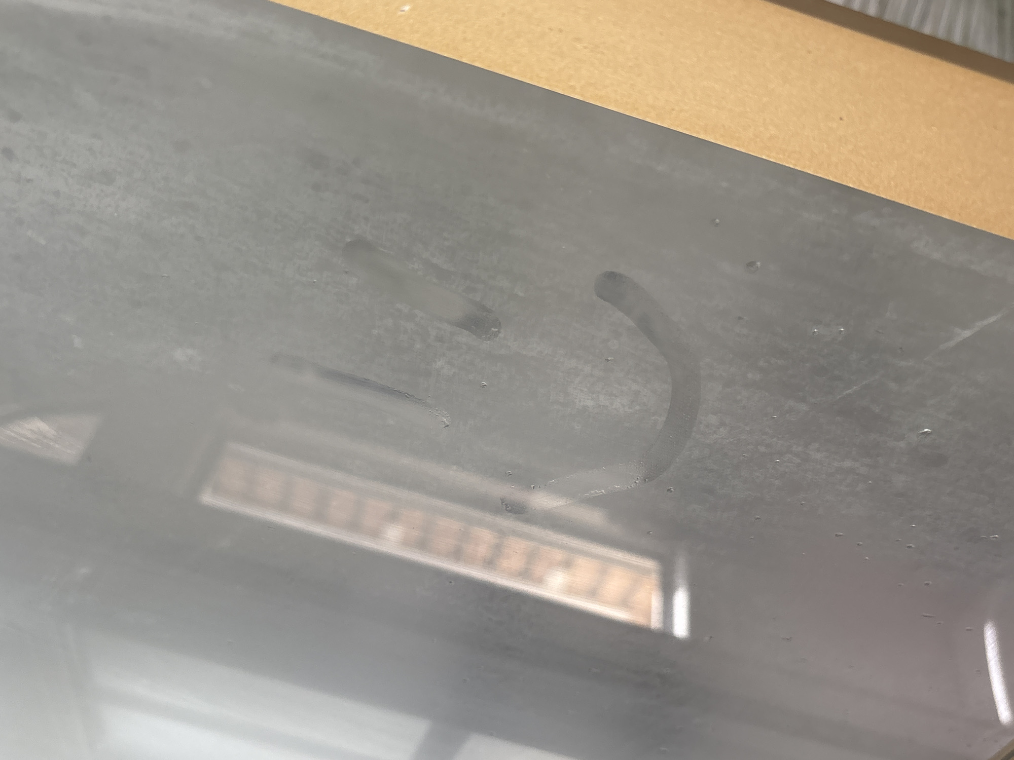 A smiley face is drawn in condensation on a glass surface, likely a window, which reflects light and a building with horizontal window panes. The condensation creates a smudged effect, obscuring details of the reflection. A portion of a tan-colored object, possibly cardboard or wood, occupies the right side of the frame. The scene appears to be indoors, with the reflection suggesting an exterior environment. There is no information in the image to suggest a specific location in Orihuela, Spain.