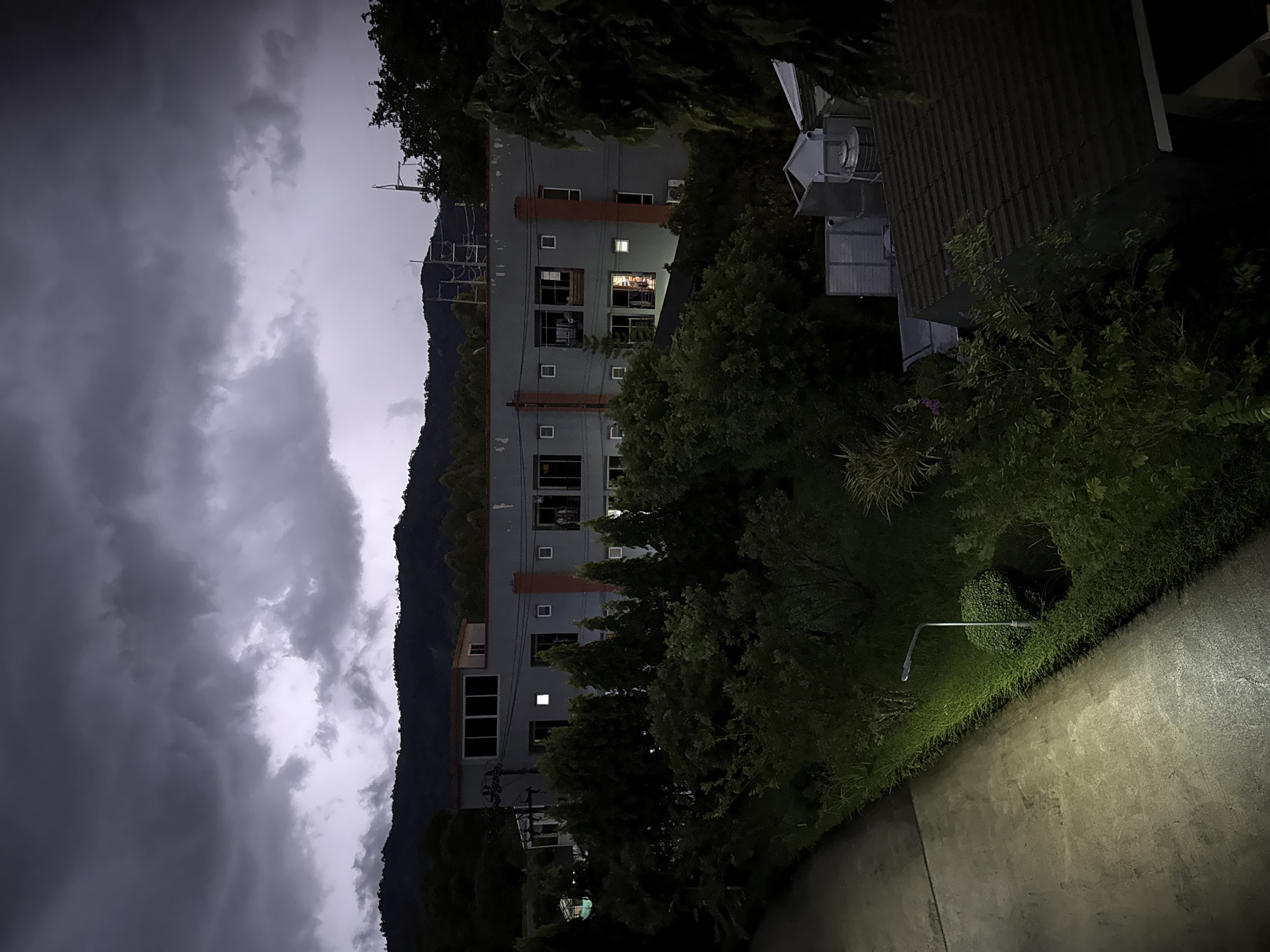 A low-angle, vertically-oriented nighttime shot shows a street in Kathu, Thailand. A gray, multi-story building with visible windows and orange accents dominates the center of the frame, partially obscured by lush trees. A dark, mountainous backdrop looms behind the building, with a stormy sky overhead. A street lamp illuminates a paved sidewalk on the right, highlighting green shrubbery and concrete. The scene evokes a quiet, overcast evening.