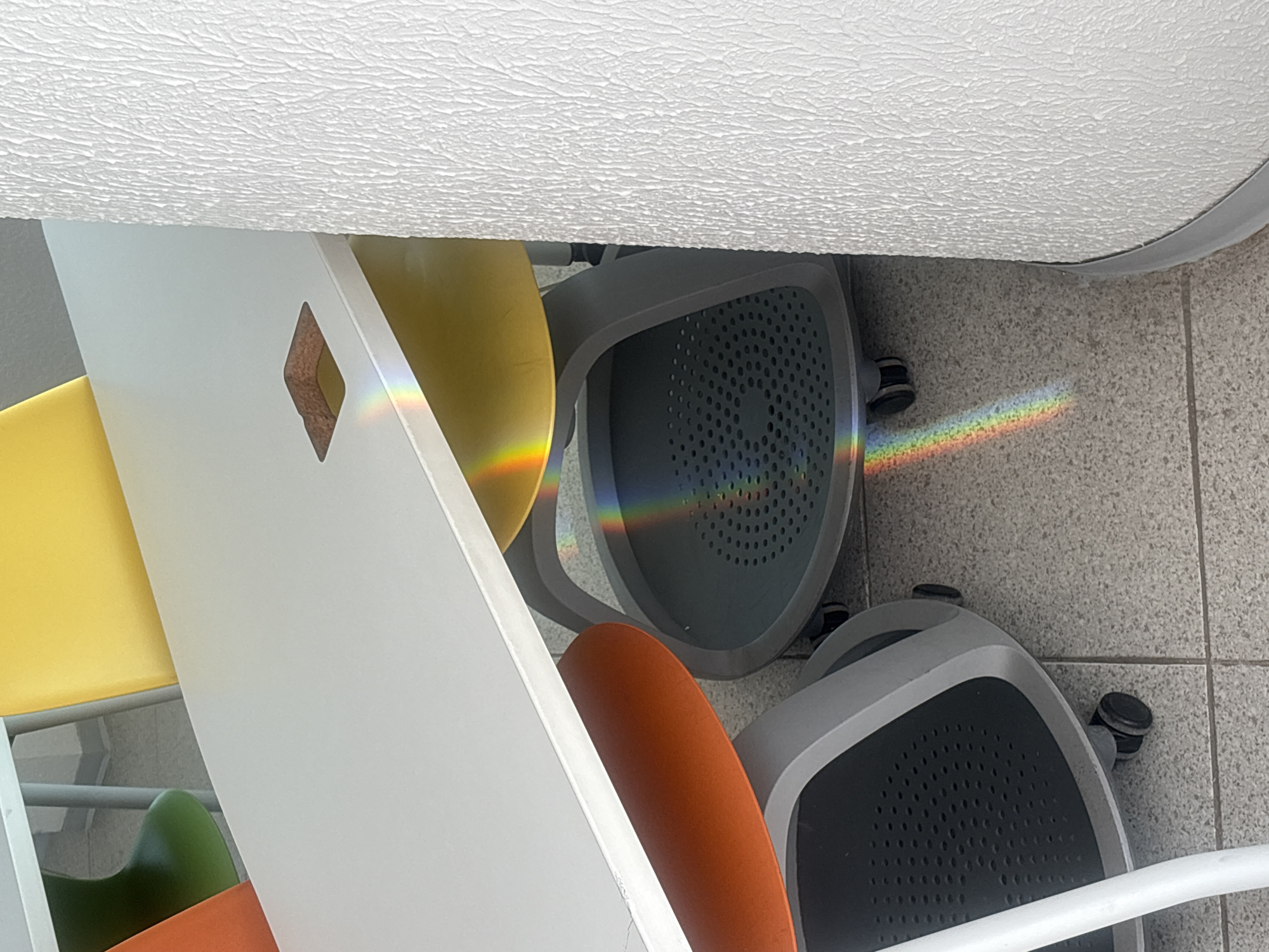 The image shows a close-up of several office chairs with a spectrum of light on the floor and chairs. The chairs are of different colors including yellow, orange, green, grey, and black. The floor is tiled in grey and the ceiling is white with a textured finish. The spectrum of light appears to be a prism effect, with the light dispersed into its constituent colors. The location in the city of Puebla, Mexico is not determinable from the image.
