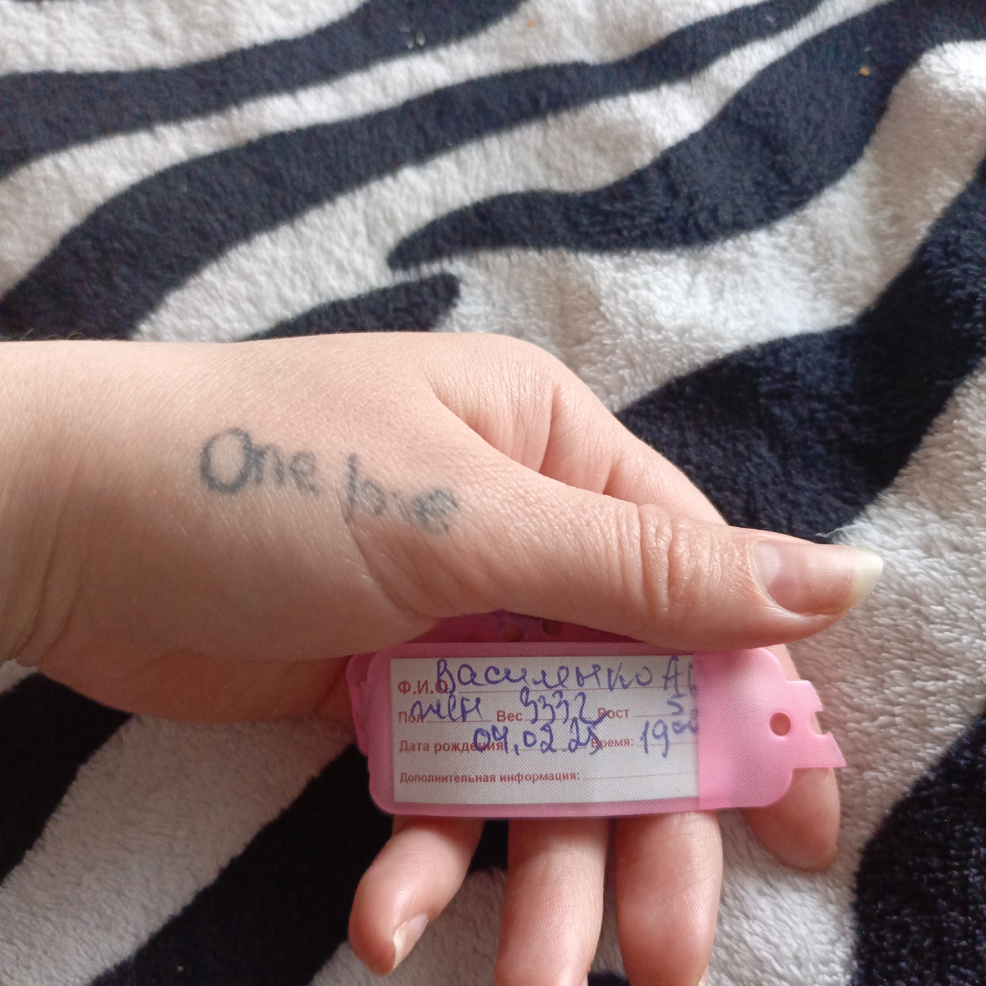 A close-up photograph shows a person's hand holding a pink identification tag, set against a background of a black and white zebra-striped textile. The person's thumb has a "One Love" tattoo. The pink tag has handwritten information written in what appears to be the Cyrillic alphabet and includes: Ф.И. Василенко AAL, вес 3332, Рост 51, дата рождения 01.02.25, Время 1908. The location and additional contextual details like the city are not discernible from the image.