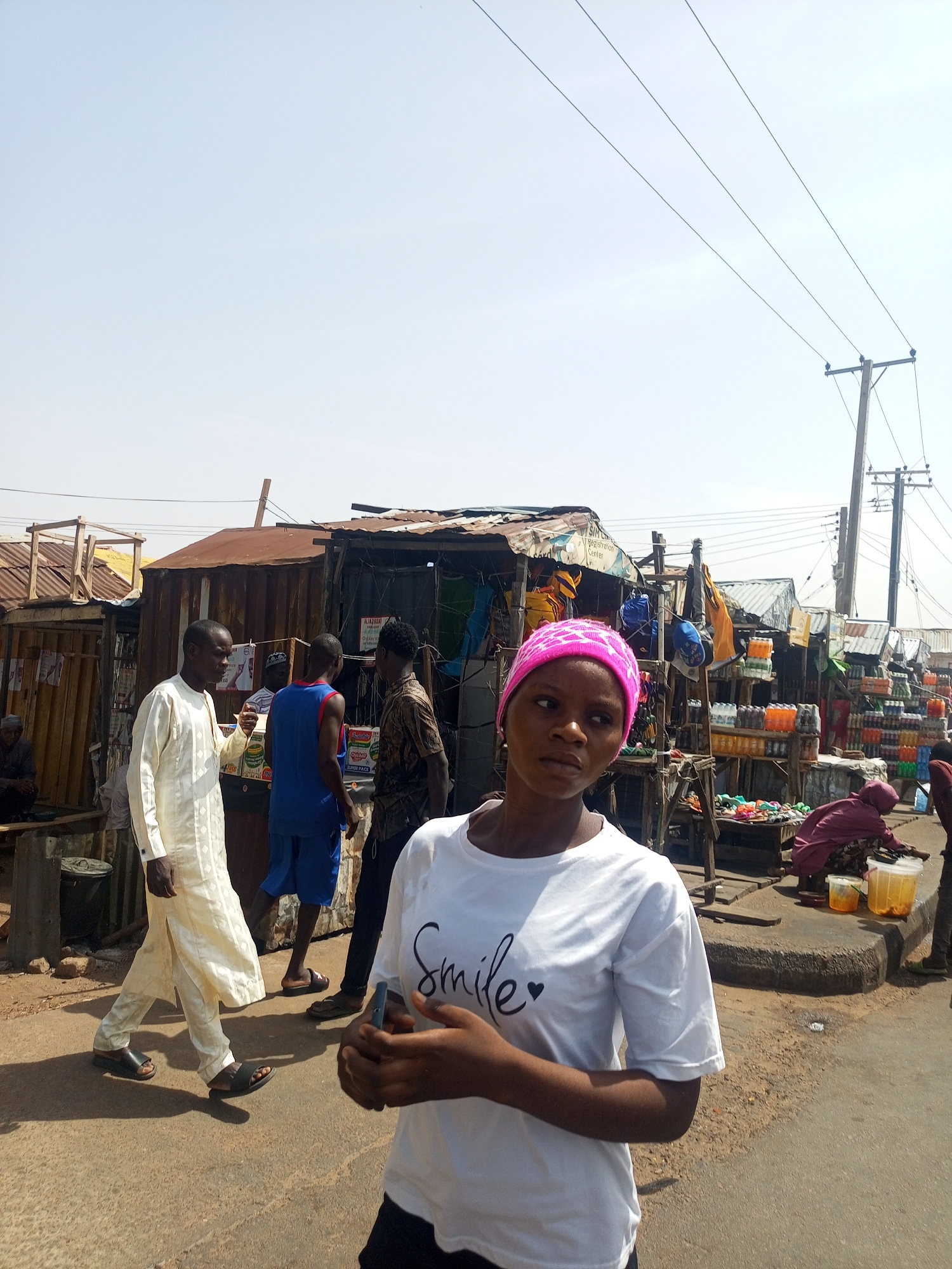 The image depicts an outdoor scene in Gombe, Nigeria, showing a street with small shops or stalls lining one side. In the foreground, a young woman wearing a white t-shirt with the word "Smile" and a pink headscarf is holding a phone. Several people are visible in the background, including a man wearing a cream-colored traditional outfit, a man in a blue tank top, and other individuals around the stalls. The stalls are selling drinks and other items. Overhead, there are power lines on wooden poles. The road surface appears to be worn, and the overall atmosphere is of a busy, bustling street scene.