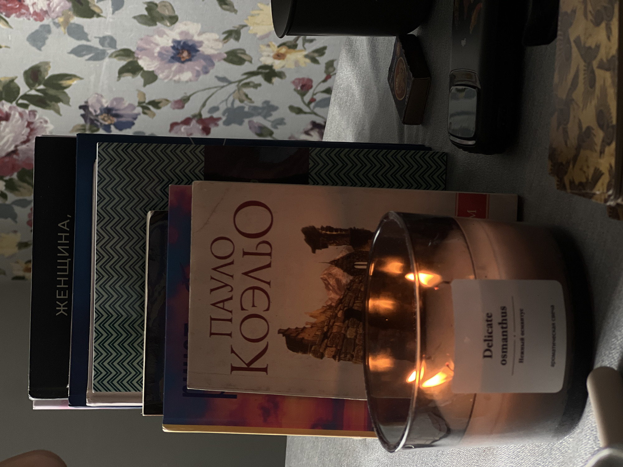 A still life scene is captured in a close-up shot, focusing on a collection of books, a lit candle, and other assorted items arranged on a surface that appears to be a table or desk. The background features a wall with floral wallpaper, depicting large, stylized flowers in shades of pink, purple, and yellow against a light blue backdrop.
In the foreground, a large glass-encased candle is lit, with visible flames casting a warm glow. A white label on the candle reads "Delicate osmanthus" in English, with smaller Russian text below it. To the left of the candle, a portion of a white object, possibly a tool or accessory, is visible.
Behind the candle, a stack of books is arranged. The topmost book has Russian text on its spine that appears to read "ЖЕНЩИНА," suggesting a title related to women. Beneath this, several other books are stacked, some with decorative patterned covers, including one with a blue and white zig-zag pattern. The spine of one book is partially visible and appears to be red. A book with a light-colored cover and the author's name "ПАУЛО" and "КОЭЛЬО" (Paulo Coelho) in large, red Cyrillic letters is prominently displayed. The cover features an illustration of a stone structure, possibly a castle or fortress, set against a mountainous backdrop with a colorful sky.
To the right of the books, a small, dark-colored object, resembling a matchbox with some visible text and imagery, is placed on the surface. Further to the right, a dark, cylindrical object, possibly a mug or a container, is visible. A dark-colored electronic device, possibly a mobile phone or a remote control, is positioned in front of the mug. At the bottom right corner, a stack of objects with a patterned surface, reminiscent of cards or a box, is visible.
The overall lighting suggests an indoor environment, likely in the late afternoon or evening, given the warm glow of the candle and the shadows cast. The scene evokes a sense of quiet contemplation or relaxation. There is no indication of the specific location in Saint Petersburg, Russia, beyond the presence of Russian text on the books.