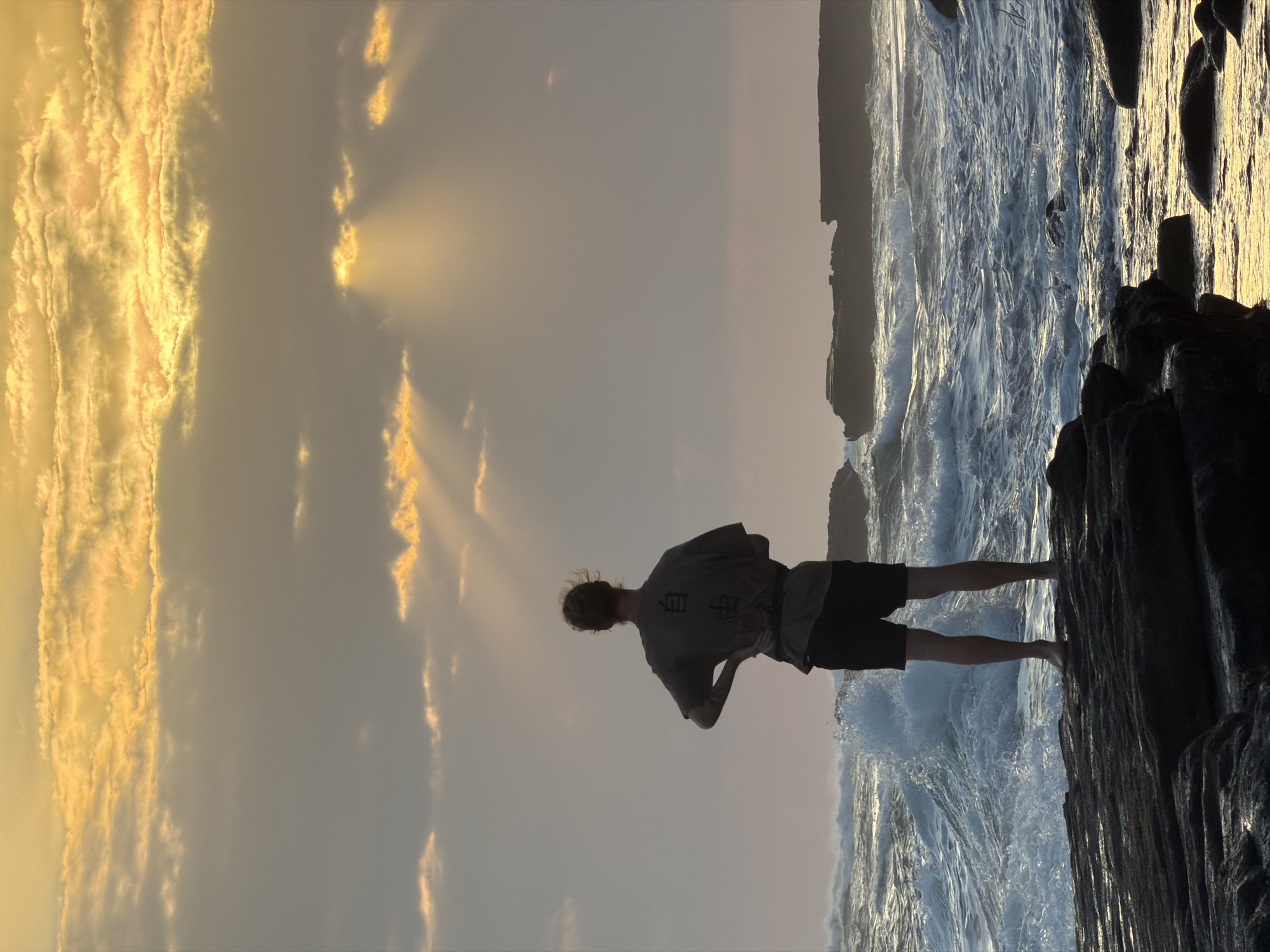 A lone person stands on dark rocks, facing a turbulent ocean at sunset. The sky above is filled with dramatic clouds, with rays of golden light breaking through, illuminating the scene. The person, viewed from behind, wears dark shorts and a t-shirt with text on the back. Their left hand is on their hip, and their hair is pulled back. The ocean is a deep blue with white waves crashing against the shore and nearby rock formations. The overall atmosphere is one of solitude and contemplation, set against the raw beauty of nature. The location is the coast of Mengwi, Indonesia.