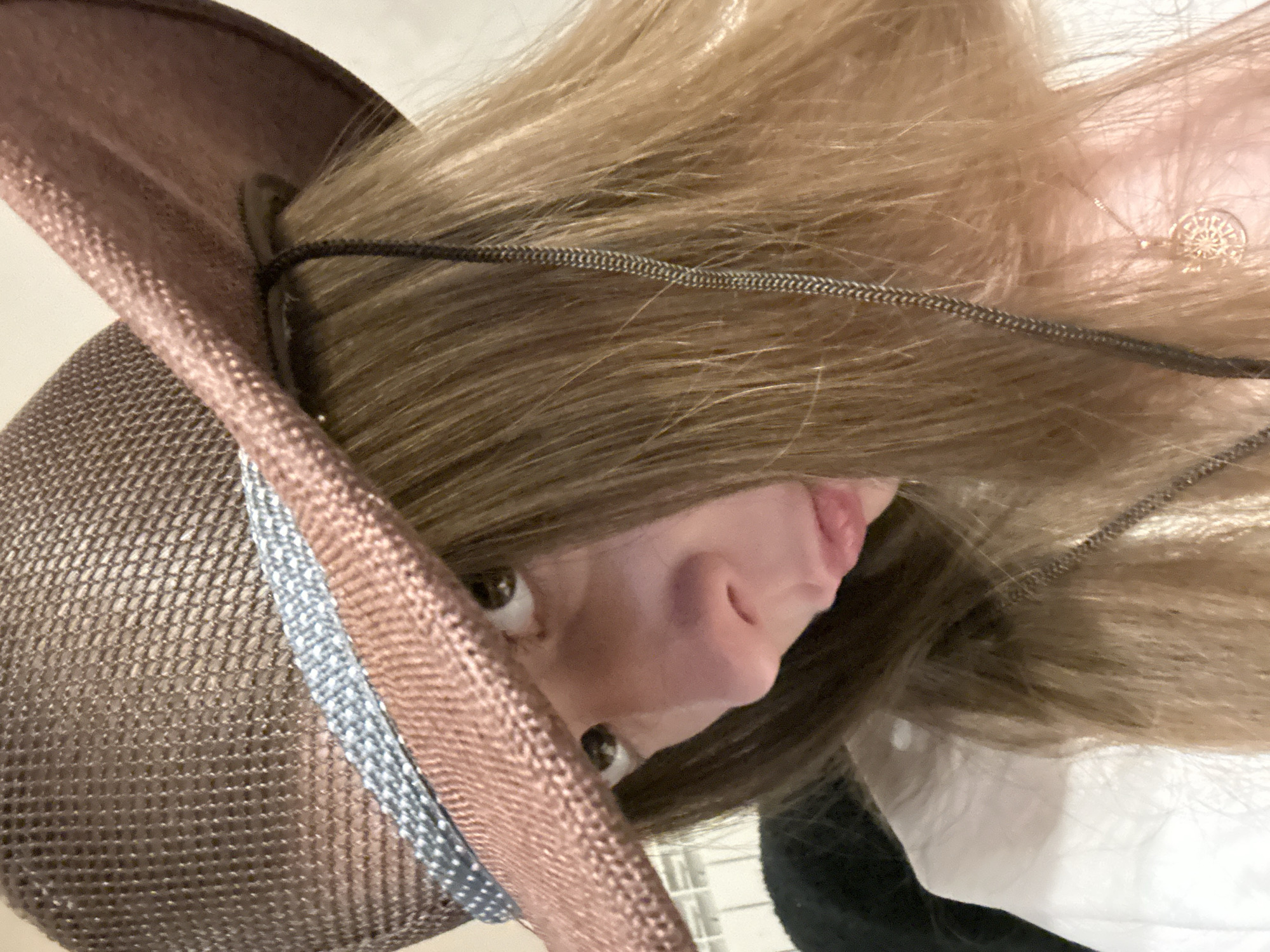A close-up, eye-level shot shows a young person wearing a brown woven cowboy hat with a blue and white patterned band and a thin cord. The hat is tilted forward, obscuring the upper part of their face, and their gaze is directed downwards and to the left. Their blonde hair, parted in the center, falls around their shoulders. They are wearing a white top and a delicate gold necklace with a circular pendant. The background is blurred and appears to be an indoor setting. The individual is located in the city of Orekhovka, Russia.