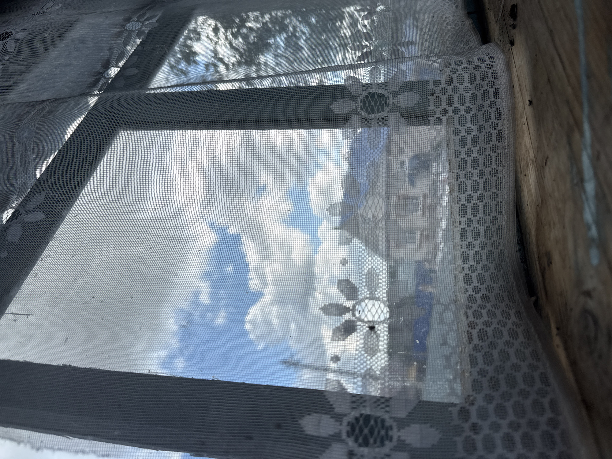 The image shows a view through a sheer lace curtain from what appears to be the interior of a building, looking out at a residential area in the city of Bolshaya Ucha, Russia. The curtain, with floral and geometric patterns, partially obscures the scene outside. Through the netting, a bright blue sky with fluffy white clouds is visible. Below the sky, there are rooftops of houses, some with visible chimneys, and trees with foliage casting shadows. The foreground consists of a wooden surface, possibly a windowsill or floor, with scattered debris. The overall impression is one of a slightly aged, rustic setting. There are no people visible in the image.