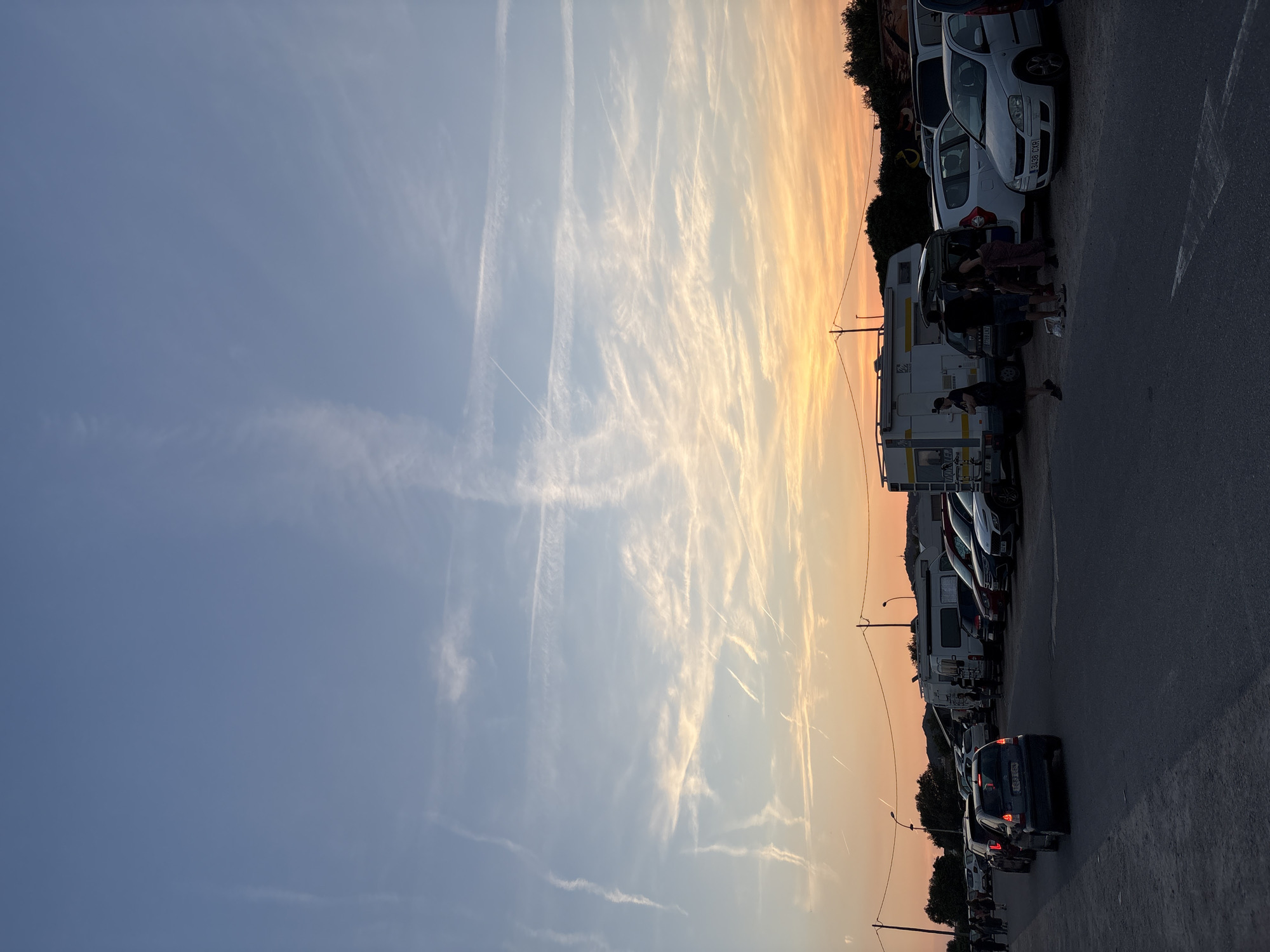 A parking lot in Cunit, Spain is lined with cars and RVs at dusk. A group of people are gathered near a white car with an open trunk. The sky transitions from blue to orange, streaked with contrails.
