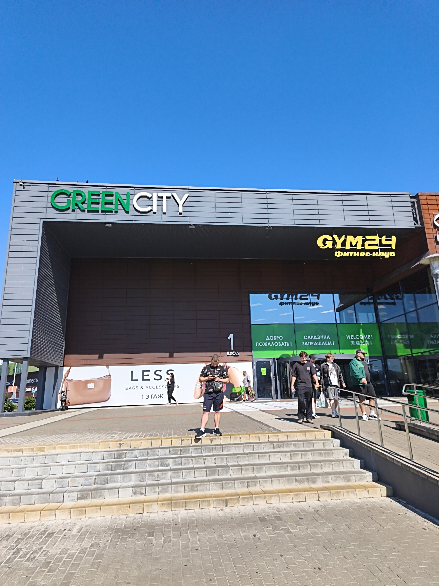The image captures the exterior of a modern shopping center, "Green City," located in Minsk, Belarus. The building features a prominent "GREEN CITY" sign in large green lettering against a gray facade. To the right, a yellow sign announces "GYM24 fitness-club" with Cyrillic text below. The entrance area is wide with glass doors and signage in multiple languages, including Russian ("ДОБРО ПОЖАЛОВАТЬ!"), Ukrainian ("САРАДЧНА ЗАПРАШАЕМ!"), and English ("WELCOME!"). A large banner displays an advertisement for "LESS BAGS & ACCESSORIES" with an image of a handbag and a scooter.
Several people are present on the stairs and walkway leading up to the entrance. In the foreground, a man wearing a black and white patterned shirt and dark shorts stands on the stairs, looking at his phone. A group of young men are walking down the stairs, one in a black t-shirt and pants, another in a white t-shirt and shorts, and a third in a green jacket and shorts. Another person in a black outfit walks across the walkway near the entrance. The environment appears to be a bright, sunny day with a clear blue sky. The ground is paved with interlocking bricks, and there are metal railings along the ramp and stairs.