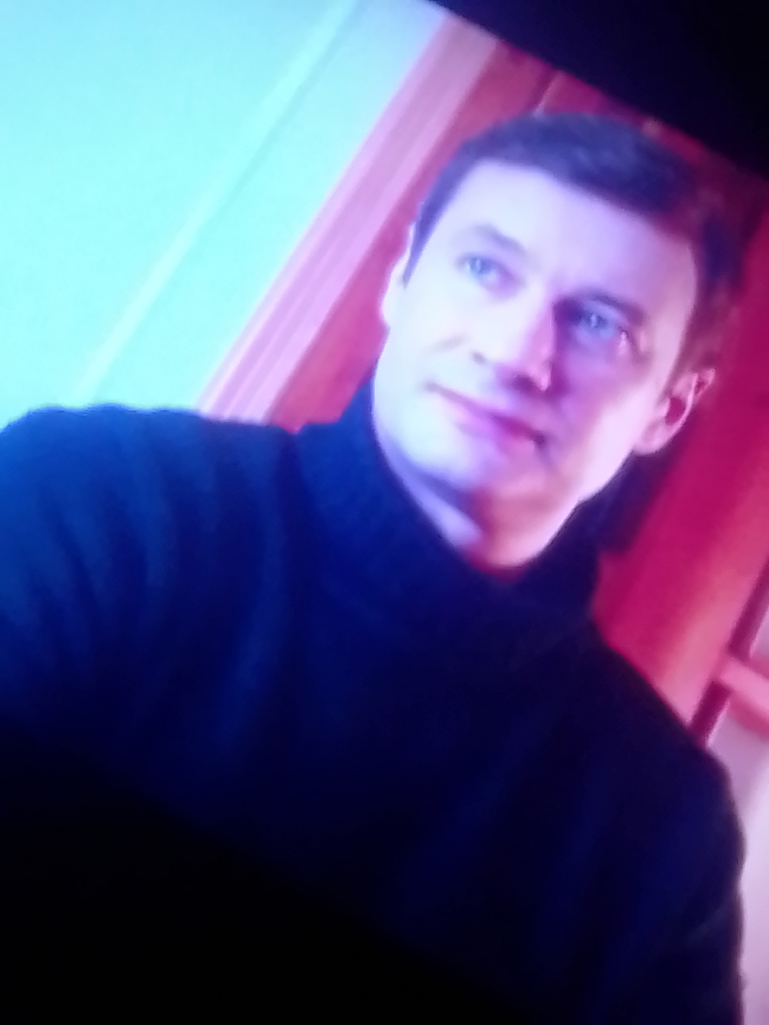 A man with short brown hair and blue eyes is pictured in a close-up, waist-up shot. He is wearing a dark turtleneck sweater. The background is out of focus and appears to be indoors with pink and blue lighting. The image is blurry and appears to be taken from a screen. There is no information available about the location in the city of Лохвицы, Russia.