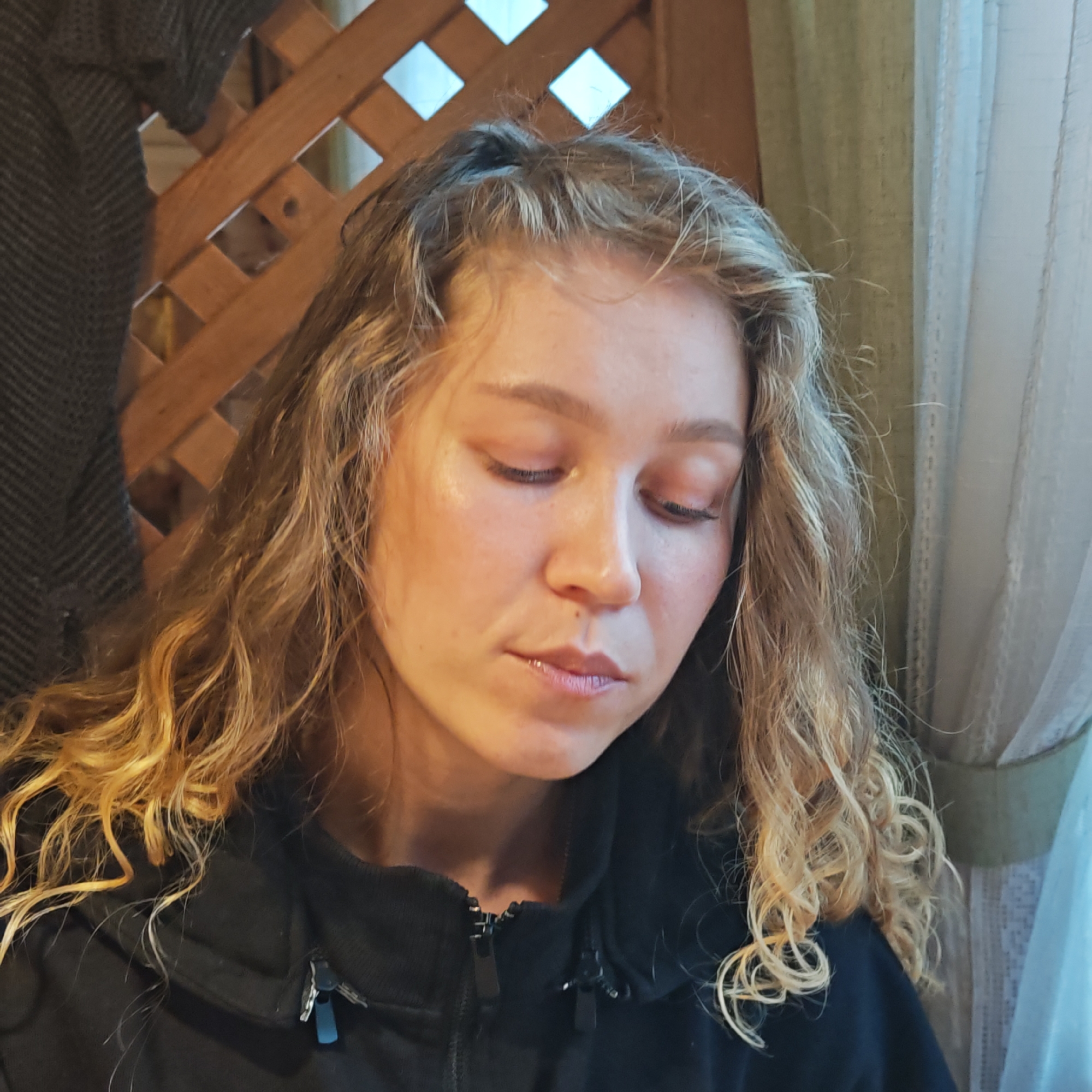 A young woman with curly blonde hair, wearing a black zip-up jacket, is looking down. She is seated indoors. In the background is a wooden lattice structure and a window with curtains. The location is suggested to be in Rostov, Russia.