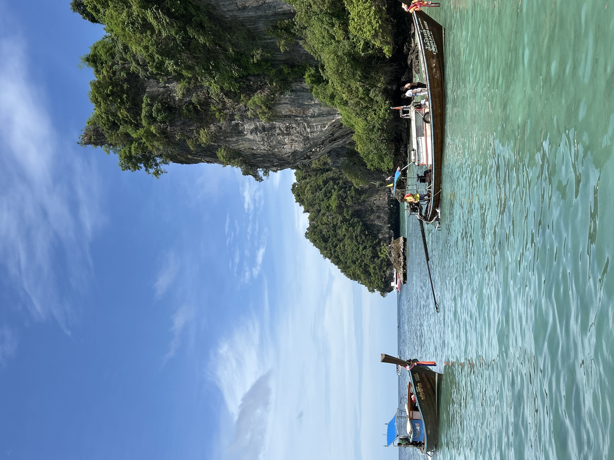 This is an eye-level, full shot of a calm, turquoise bay near a sheer, tree-covered limestone cliff face in Ko, Thailand. Two longtail boats are docked in the bay. The boat on the left is brown with a blue canopy and a Thai flag. The boat on the right is also brown and has several people on it, including a person in an orange life vest. The water is clear and ripples gently. The sky is a bright blue with scattered white clouds.