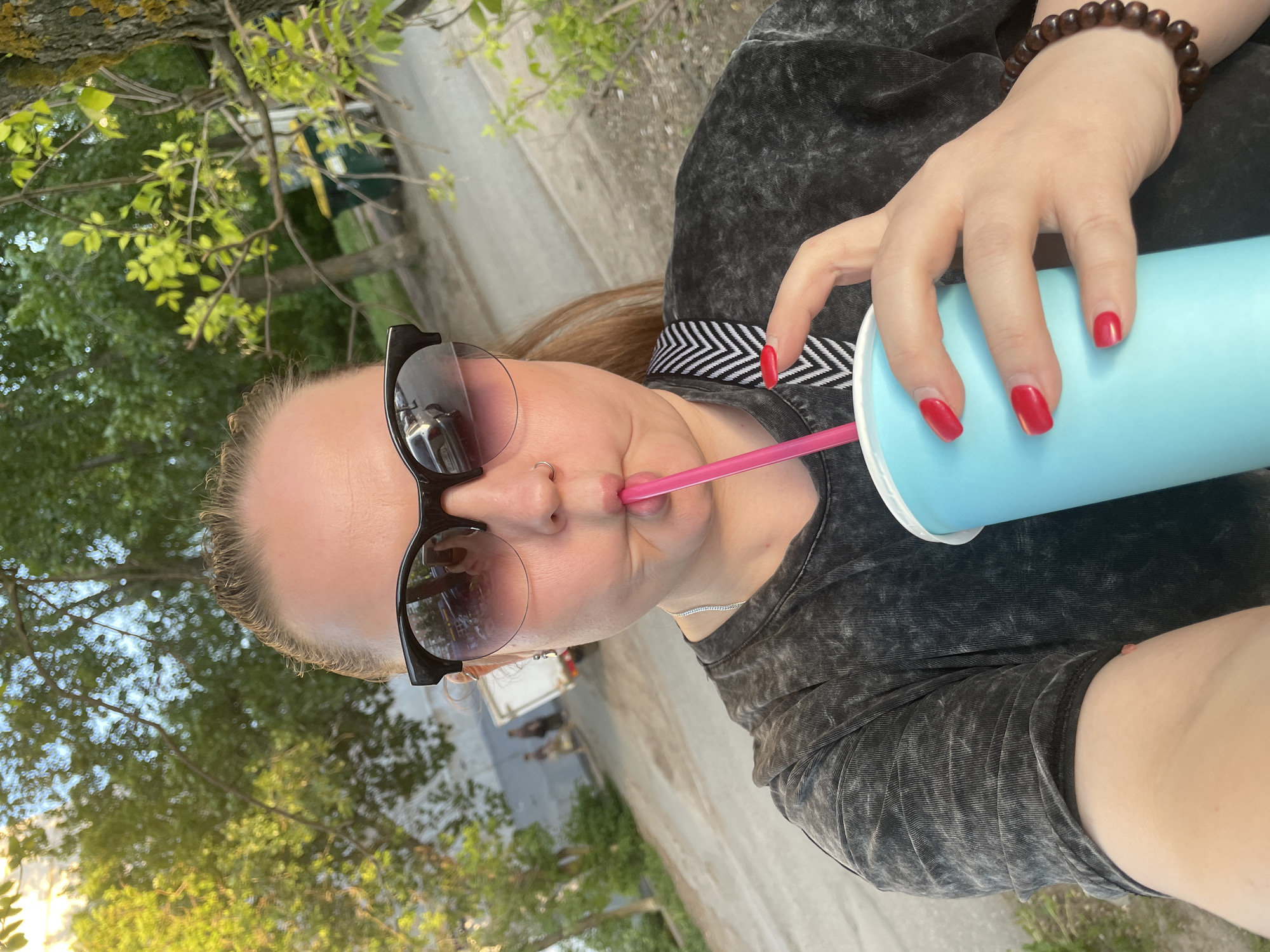 A woman with sunglasses is drinking from a light blue cup with a pink straw. She wears a dark gray shirt and has red nail polish. A bracelet is visible on her right wrist. The background includes trees and a blurred cityscape. Although it is not possible to verify the location as Izhevsk, Russia, based solely on this image, the background appears to be an urban or suburban environment.
