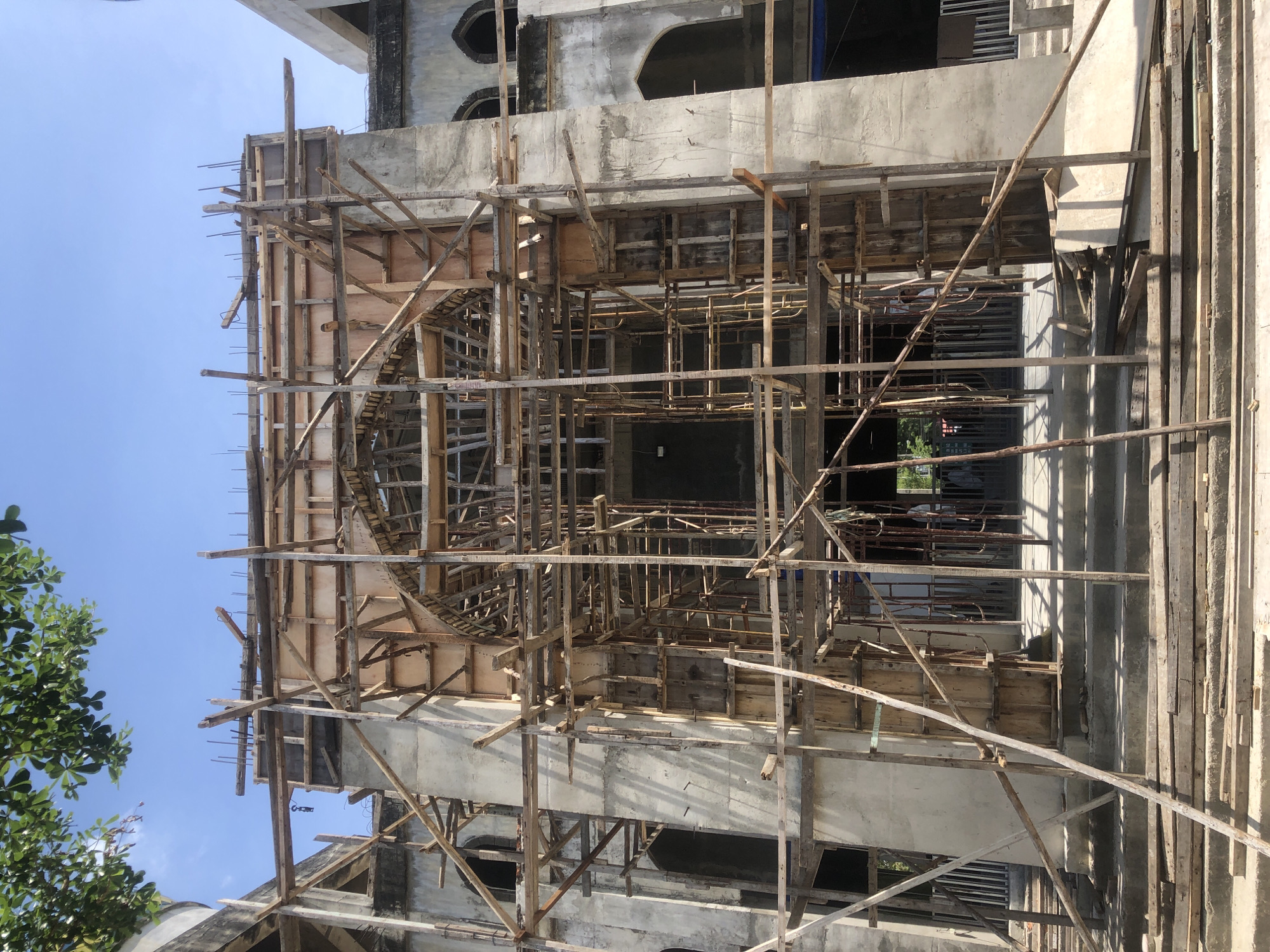 The image shows the exterior of a building under construction in Lhokseumawe, Indonesia. The structure appears to be a grand entrance, possibly for a public building or mosque, with a large arched opening. The construction is in an advanced stage, with concrete walls and arched windows visible. A significant amount of wooden scaffolding surrounds the entrance, indicating ongoing work on the facade and upper levels. Several wooden planks are laid out in front of the entrance, likely for temporary walkways or material staging. The sky is clear and blue, suggesting it is a sunny day. The overall scene depicts a bustling construction site.