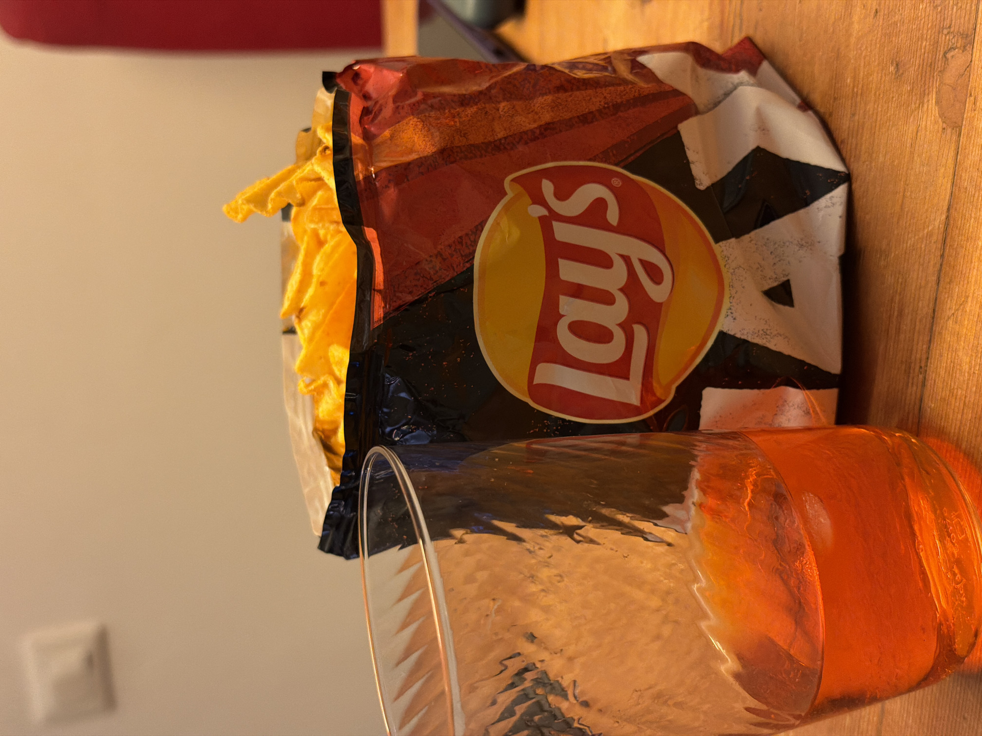A bag of Lay's potato chips is partially open, revealing chips inside. Next to it is a glass containing a pale orange liquid. The scene appears to be on a wooden surface indoors. No people are visible. The location is unspecified and cannot be determined from the image alone. Therefore, it is unknown if the image was taken in Milan, Italy.