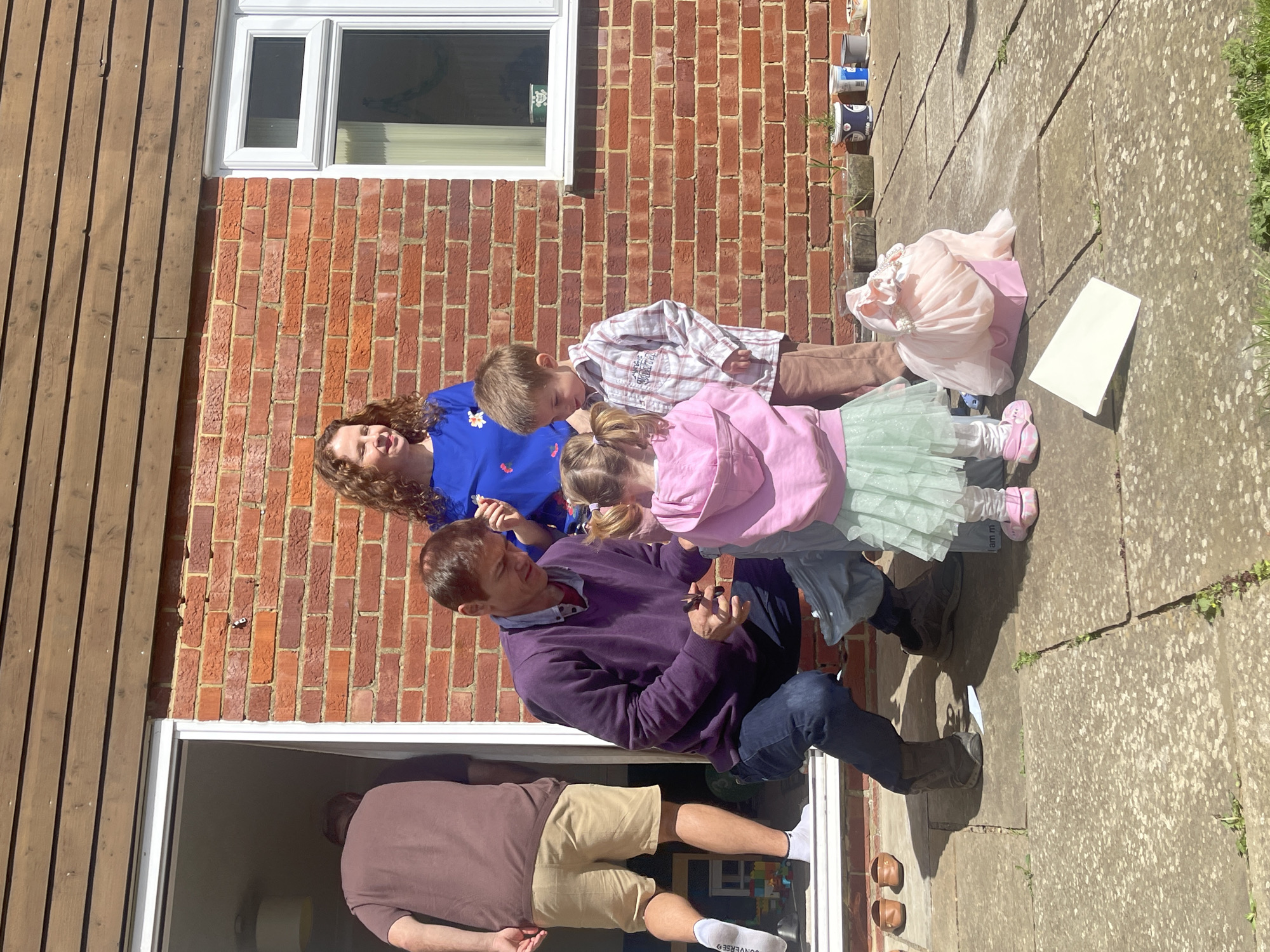 The image depicts a family gathering outdoors, likely a garden patio adjacent to a brick house with a wooden-paneled exterior section and a white-framed window.
There are three adults and two children visible. One adult, a woman with curly hair wearing a blue dress, stands to the side of the brick wall. A man in a purple sweater and jeans is kneeling, apparently interacting with two children, a young boy in a plaid shirt and a young girl in a pink hooded jacket and a tutu skirt. A third adult wearing a brown shirt and khaki shorts stands in the doorway of the house.
A pink shopping bag and a white rectangular object are on the paved area. Some potted plants are seen affixed to the brick wall.
I lack enough information to confirm this image was taken in Maidstone, United Kingdom.