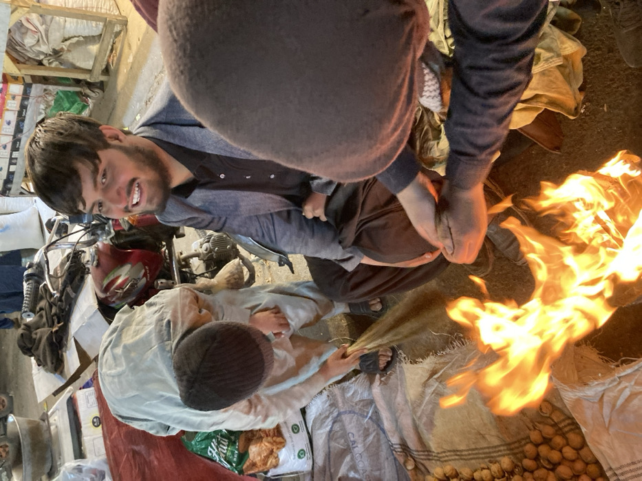 A group of men is gathered around a fire. One man looks at the camera with a smile. Another man is using a broom to tend the flames. Bags filled with produce sit nearby, with a motorcycle and other items surrounding the group. The scene appears to be taking place in an outdoor market or similar setting.