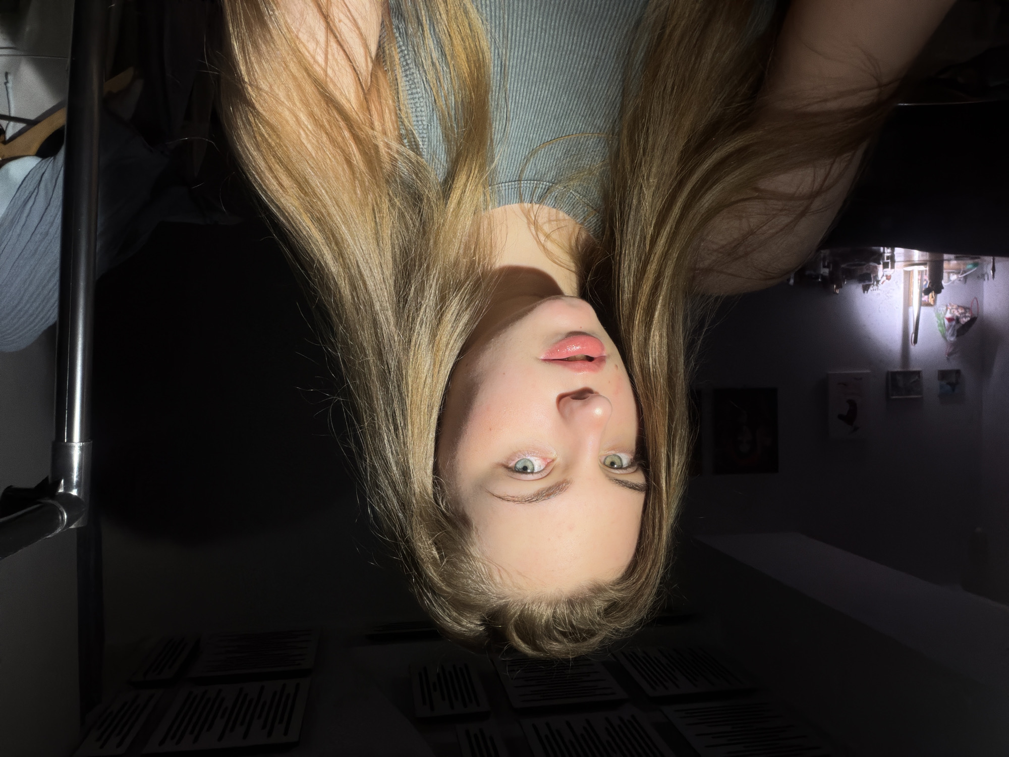 The image shows a close-up of a young woman with long, blonde hair. She has light-colored eyes and is wearing a grey tank top. The lighting suggests she is indoors, possibly in a room with a dark background and some visible objects like a desk with a lamp and some artwork on the wall. There is no indication of the location being in Dnipro, Ukraine.