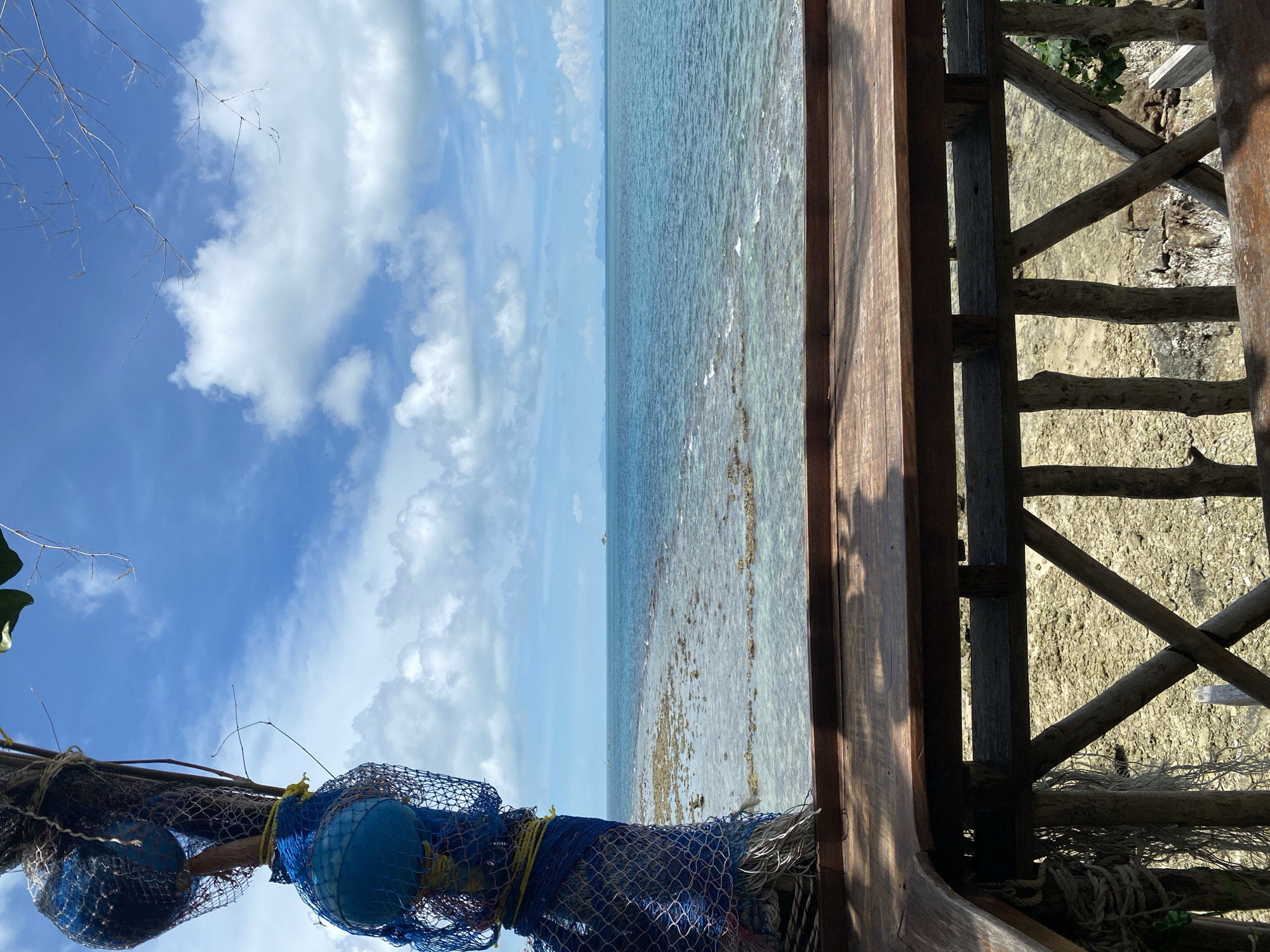 The image depicts a tropical coastal scene viewed from a wooden deck. In the foreground, a weathered wooden table and railing are visible, with rough-hewn wooden posts supporting the structure. To the left, a rough pole adorned with blue netting and spherical objects, possibly buoys, hangs suspended. Beyond the deck, the turquoise ocean stretches to the horizon, dotted with faint islands. The water near the shore is shallow and clear, revealing a rocky or coral seabed. The sky above is a vibrant blue, punctuated by scattered white clouds. The overall impression is one of a relaxed, outdoor setting by the sea. The location is identified as Ko Pha Ngan, Thailand. There are no people visible in the image.