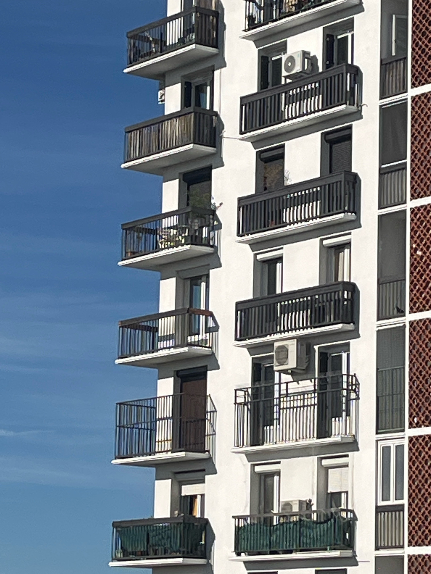 A side view of a modern apartment building shows a white facade with multiple balconies, each with dark railings and light trim. The balconies are stacked vertically. Some balconies contain outdoor furniture, such as chairs and a small table, while others are sparsely decorated or contain air conditioning units. On the right side of the building, a patterned section of reddish-brown brickwork is visible. The sky is a clear, bright blue.