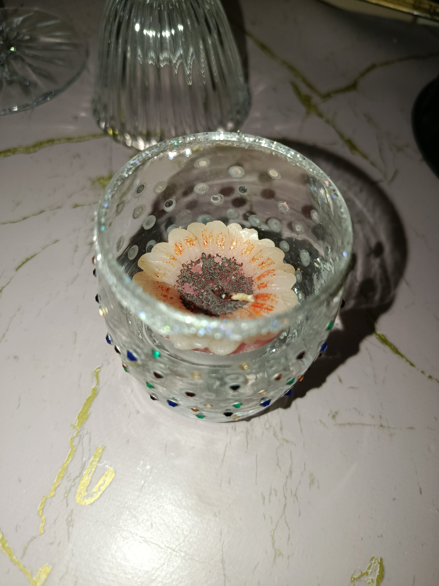A close-up shot captures a decorative glass votive holder with a flower-shaped candle inside. The votive holder is adorned with small, colorful gems along its rim and a textured pattern of raised dots on its surface. The candle, shaped like a daisy or sunflower, has white petals with orange accents and a dark, textured center. The votive holder sits on a light-colored surface with subtle veining and streaks of gold paint, resembling a marble countertop. In the background, two clear glass objects, possibly wine glasses or decanters, are visible, one with vertical fluting. The lighting creates soft shadows and highlights on the objects and the surface. The image composition focuses on the votive holder, with a shallow depth of field blurring the background elements. There is no indication of location in Tashkent, Uzbekistan.