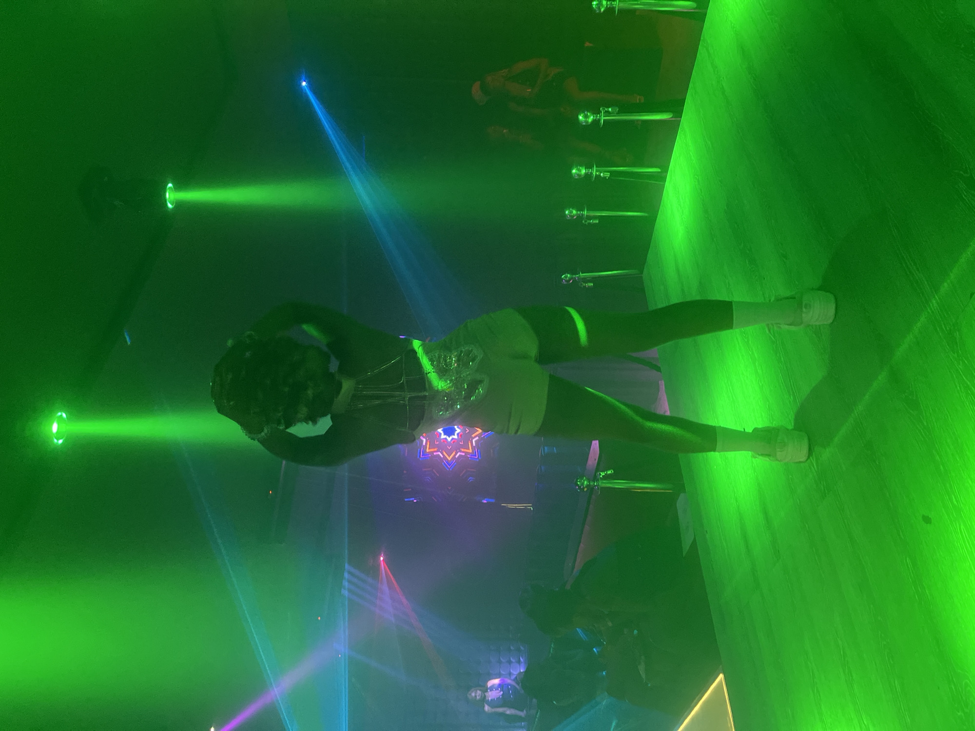 In a dimly lit club environment, bathed in vibrant green and blue laser lights, a person is performing on a raised platform. They are dressed in a sparkly top and white shorts, with white socks and sneakers. Their pose suggests a dance or acrobatic performance, with arms raised and legs spread wide, creating a dynamic silhouette against the luminous atmosphere.
In the background, other individuals can be seen scattered throughout the club, some appearing to be seated or standing at a bar, while others are blurred figures adding to the ambiance. Several vertical poles with decorative tops are visible on the platform, possibly part of the stage setup or for aerial performance.
The scene is dominated by the intense green glow of the stage lighting, with streaks of blue and purple light cutting through the haze, indicating the presence of fog machines or smoke. A decorative star-shaped motif is visible on the back wall, adding to the visual spectacle. The overall impression is one of a lively nightlife setting, likely a club or entertainment venue in Howayi, Nigeria, where a performance is captivating the audience. The time of day is evening or night, consistent with a club setting.