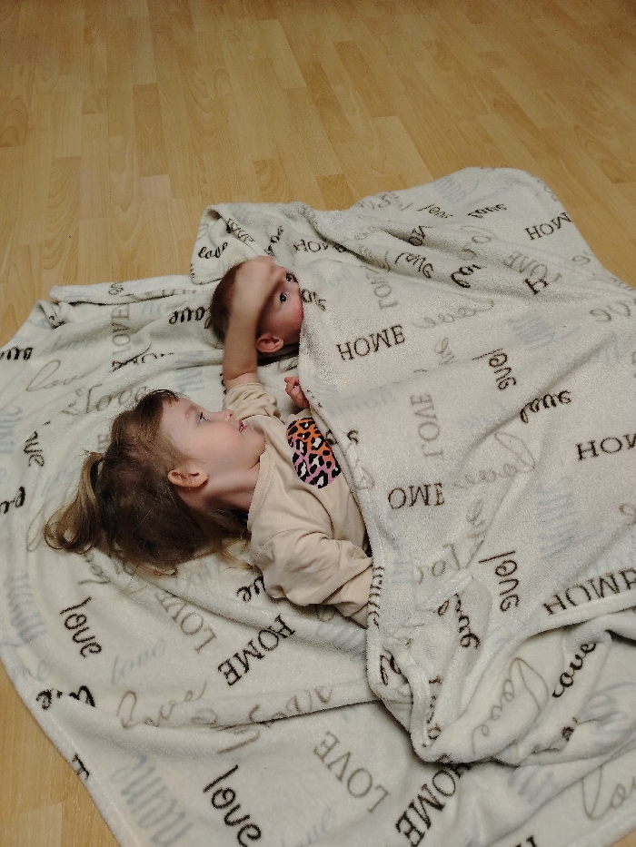 The image captures a heartwarming moment between two young children playing or relaxing on a light-colored wooden floor. Both children are partially covered by soft, cream-colored blankets that feature a repeating pattern of words like "LOVE" and "HOME" in various orientations and fonts (both block letters and a script style).
On the right side of the frame, a baby or very young toddler is mostly nestled under one of the blankets. Their head, with light brown hair, and part of one arm are visible. This child's blue eyes are wide and focused upwards and slightly to the left, conveying a sense of curiosity or engagement. Their hand is raised near their face.
Adjacent to this child, on the left, lies a slightly older toddler. This child has light brown hair pulled back into a small ponytail and is wearing a long-sleeved, light-beige top with a vibrant, multicolored animal print (appearing to be a leopard or cheetah print) heart graphic on the chest. This toddler is positioned looking towards the younger child, suggesting interaction or observation.
The scene takes place indoors, indicated by the smooth wooden flooring. The atmosphere appears calm and playful, with the children seemingly enjoying their time together under the cozy blankets. The words "LOVE" and "HOME" are distinctly visible multiple times on both blankets.