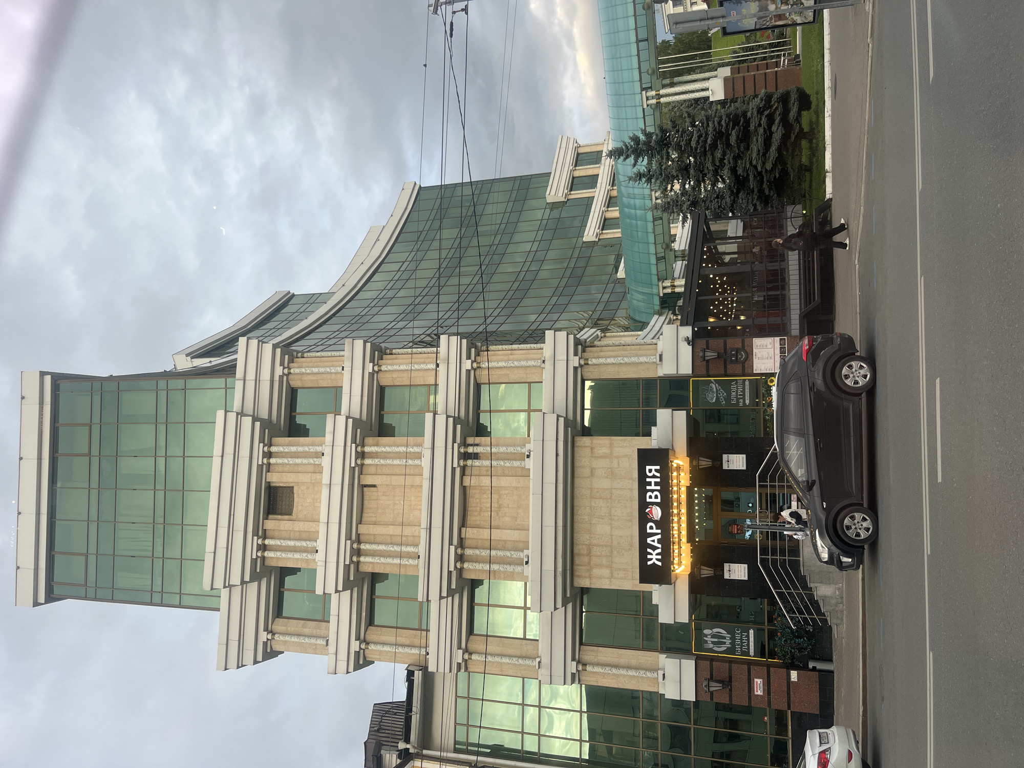 A multi-story building with a curved glass facade and a rectangular section adorned with columns is visible from a street in Kazan, Russia. The building features green tinted windows and architectural elements resembling classical columns. A sign with Cyrillic text, "ЖАРОВНЯ" (Zharovnya), is illuminated above the entrance. A dark SUV is parked on the street in front of the building, and a person is standing near the entrance. Adjacent to the building is an outdoor seating area with string lights. Several evergreen trees are planted near the structure. The sky is overcast.