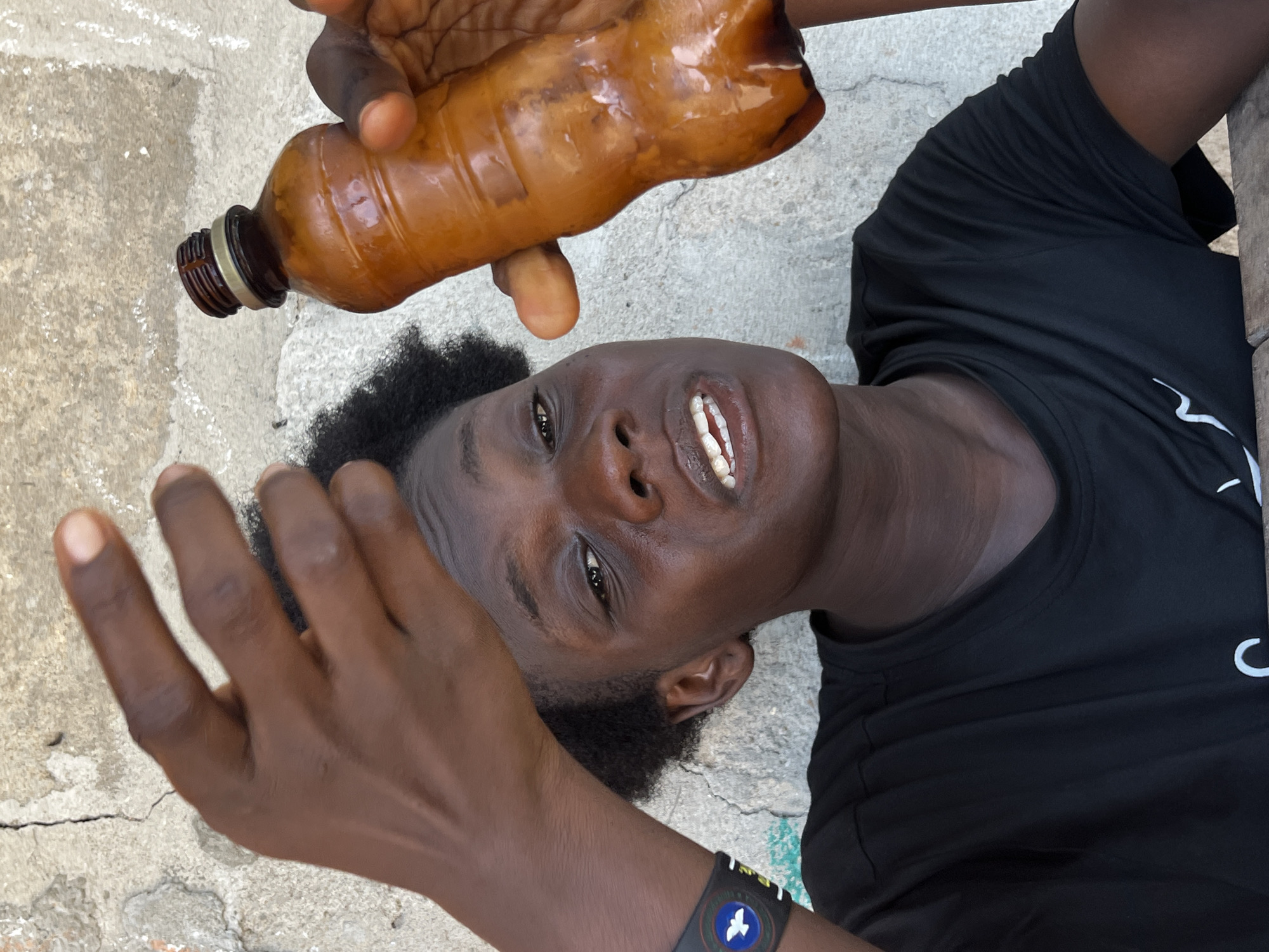 A dark-skinned person is holding a brown plastic bottle in their right hand above their head, looking directly at the camera. The bottle appears to be filled with a dark liquid. The person is wearing a black t-shirt and a wristband with a bird design on their left wrist. The background consists of a cracked concrete surface. The image was taken in Badagry, Nigeria.