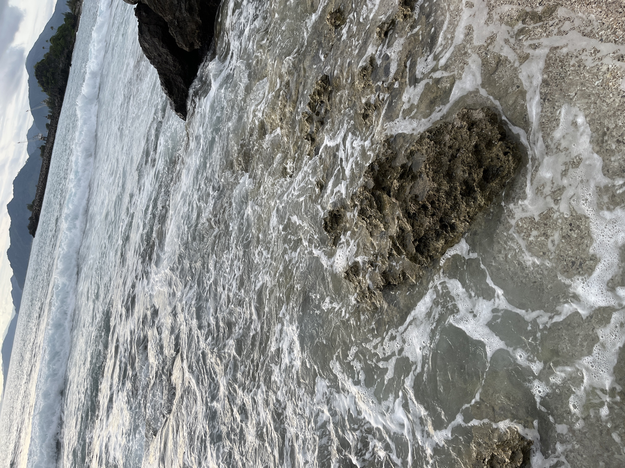 The image shows a rocky shoreline with waves gently washing over the stones. The water is clear, allowing the seabed to be visible in some areas. The rocks are dark and rough, with some covered in algae or seaweed. The waves are creating a foamy white pattern as they recede from the shore. In the background, a dark, imposing rock formation juts out of the water, casting a shadow on the wet sand. The scene suggests a natural, undisturbed coastal environment. The lighting appears to be natural, possibly indicating daytime, though the overcast sky hints at a cloudy day. The overall mood is serene and tranquil, evoking the peacefulness of the ocean. There is no visible text in the image.