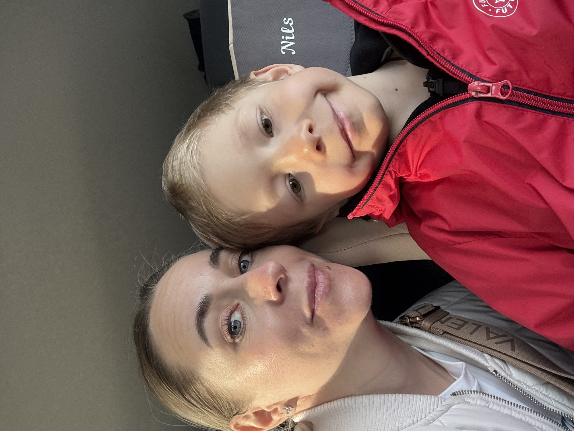 A woman and a young boy are in a vehicle. The woman has light skin, light hair pulled back, and is wearing a white jacket with a "VALENTINO" strap. The boy has light skin and light hair. He is wearing a red jacket. The vehicle is likely in the city of Chaikovsky, Russia. The image is oriented vertically.