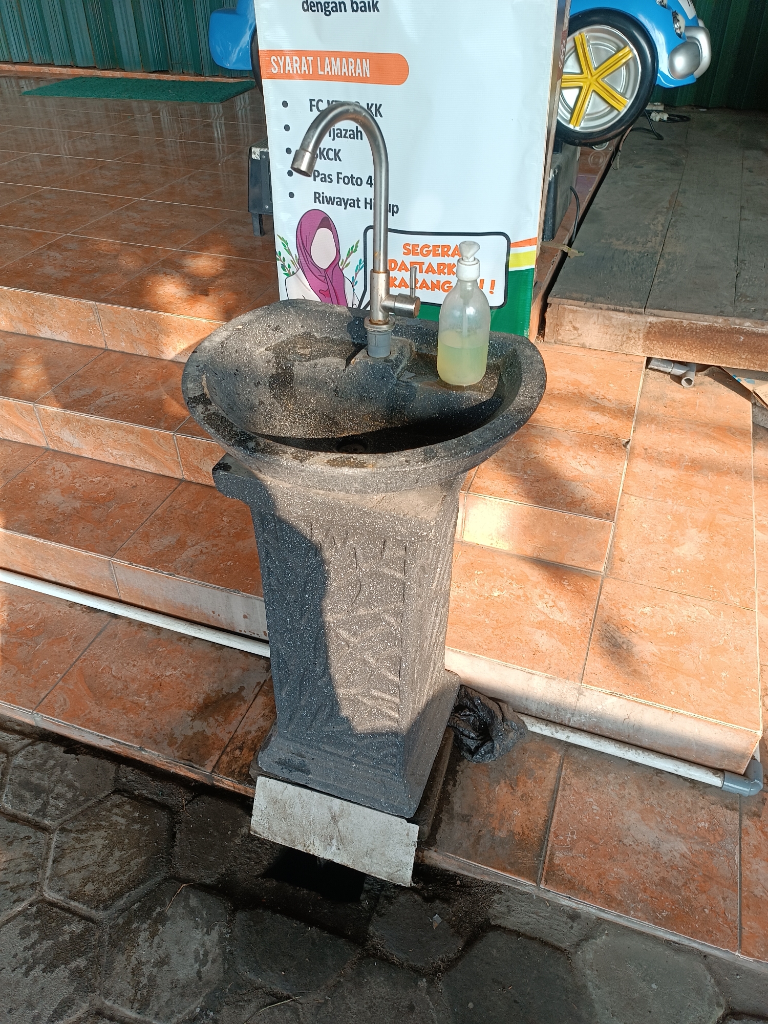 An outdoor handwashing station is situated on a tiled patio in front of a building. The station consists of a dark gray, textured pedestal sink with a chrome faucet. To the right of the faucet, a clear plastic bottle filled with light yellow liquid, presumably hand sanitizer or soap, is placed on the edge of the sink. A white sign with Indonesian text is visible behind the sink; it lists application requirements and encourages immediate registration. The ground is covered with reddish-brown tiles, and a section of dark, hexagonal paving stones is present in the foreground, showing signs of being wet. The scene is illuminated by sunlight, casting shadows. A small black plastic bag is on the ground near the base of the sink. In the background, a blue object, possibly a toy or decorative item with a yellow wheel, is partially visible. The location is identified as Imogiri, Indonesia.