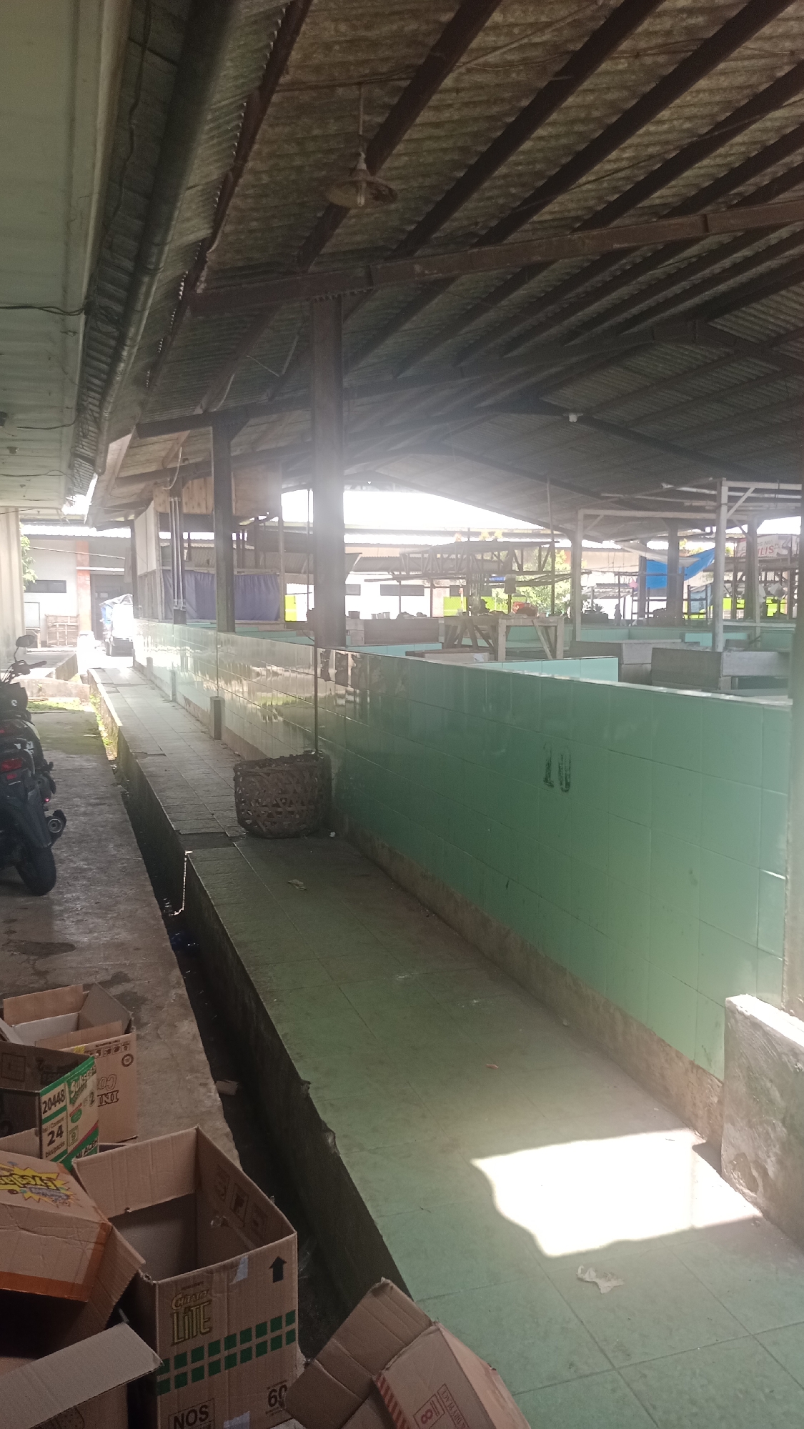 The image depicts an open-air market stall area in Cogreg, Indonesia. The structure is covered by a corrugated metal roof supported by metal beams and wooden posts. Green tiled walls line the market stalls, some of which appear to be empty. In the foreground, several cardboard boxes are piled on a concrete walkway. To the left, a motorcycle is parked. Sunlight streams through the roof, creating bright patches on the floor and illuminating some of the stalls. The scene appears to be a functional, albeit somewhat weathered, market setting.
